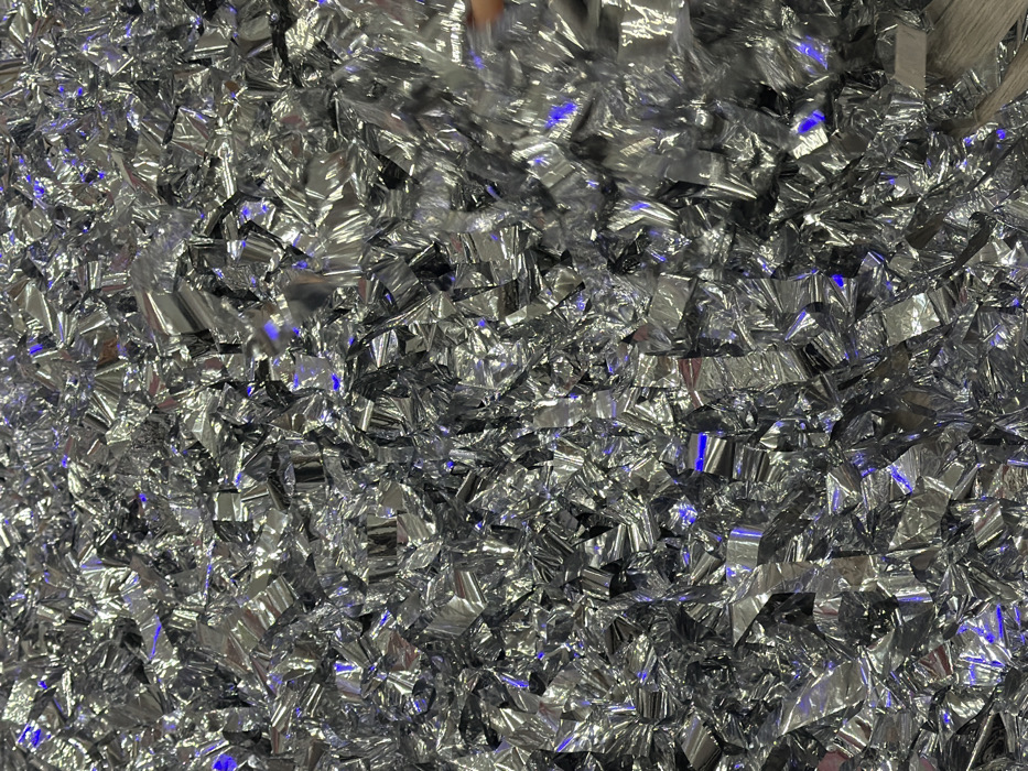 A dense, reflective field of small, silver, confetti-like pieces fills the frame. The light reflecting off the metallic material creates a shimmering, textured effect, with varying shades of gray and white. Scattered blue light adds depth to the scene. The fragments are randomly oriented, creating a dynamic and visually captivating surface.