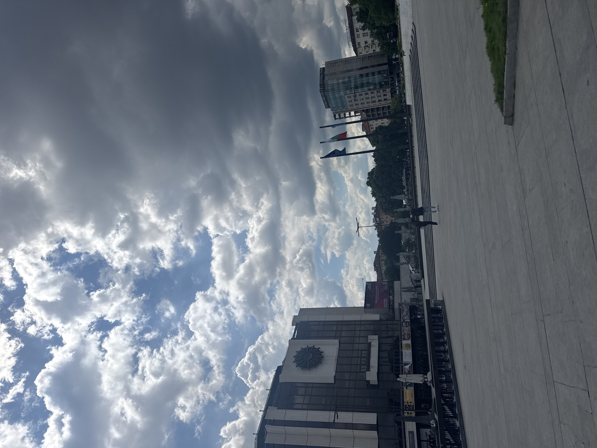 The image shows a view in Sofia, Bulgaria, near the Largo complex. The sky is filled with dramatic clouds, with patches of blue sky visible. Buildings are visible on both sides of the frame. On the left, there is a building with a large sun-like sculpture on its facade and various advertisements. On the right, there is a modern hotel building with flags in front of it. A wide concrete pedestrian area extends from the bottom of the frame towards the horizon, with some people walking. There is a grass verge on the far right side.