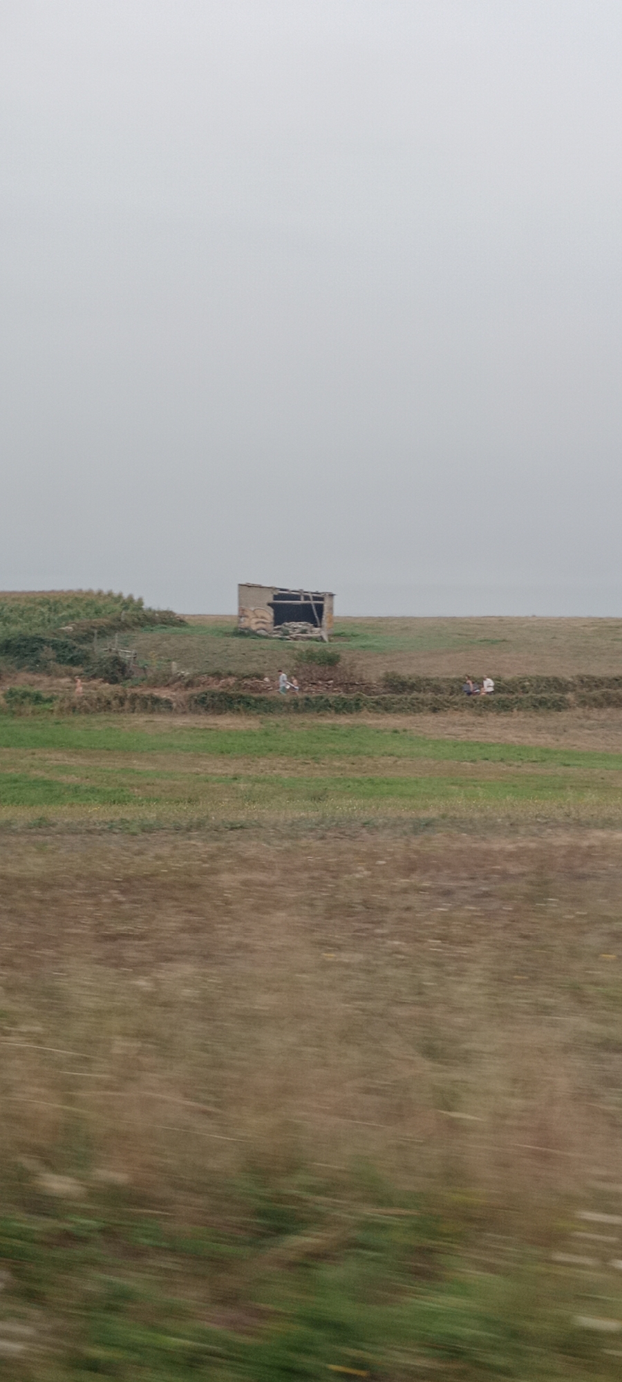 A wide shot shows a desolate landscape under an overcast sky. In the middle ground, a dilapidated, partially roofless structure sits on a grassy knoll. Faded graffiti adorns the exterior of the building. In front of the structure, two figures, possibly a child and an adult, are walking. To the left of the structure, another figure can be seen further in the distance. The foreground is a blur of brown and green, suggesting motion. The scene appears to be in a rural area, possibly A Rochela, Spain, given the agricultural fields and the rustic nature of the building.