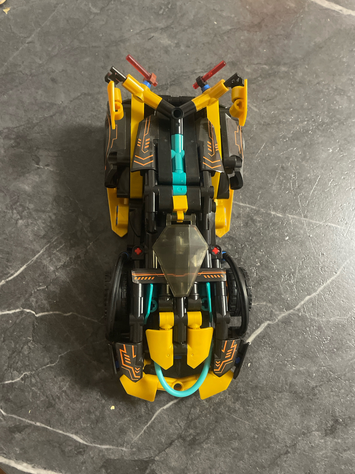 An overhead, eye-level shot captures a yellow and black Lego race car model resting on a dark gray, marbled surface. The car is oriented vertically, with its front facing upwards. The model features prominent yellow body panels, accented with black and orange detailing. A transparent cockpit canopy sits towards the front of the car. A light blue tube-like piece curves around the underside of the rear of the vehicle. The background consists of the same marbled surface, with no other objects or people visible. The image does not provide any information about the city of Baranovka, Russia.