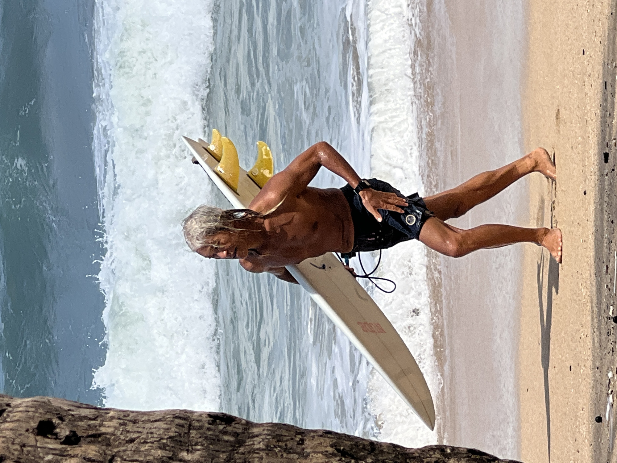 A shirtless, bare-chested man with long, graying hair is walking on the beach at Kuta, Indonesia, carrying a surfboard. He is wearing dark swim trunks and a watch on his left wrist. The surfboard is light-colored with a red logo and yellow fins. The ocean behind him has significant waves breaking, with white foam and blue water. A portion of a tree trunk is visible on the left side of the frame. The sand on the beach appears wet near the waterline and dry further up.