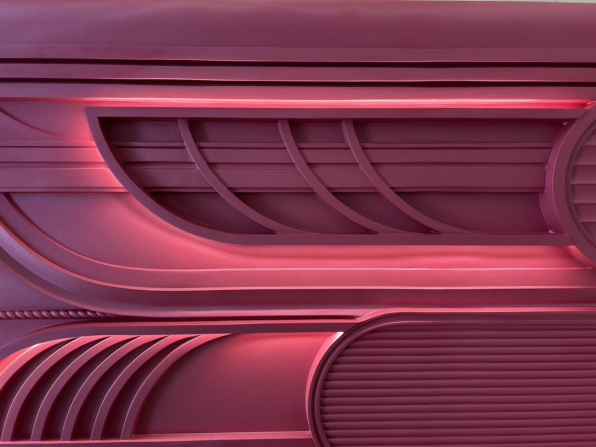 This media file presents a detailed, abstract view of an architectural or decorative panel, rendered in a rich maroon or deep pink color. No people or specific events are depicted. The panel features a complex, three-dimensional design composed of various geometric and organic patterns.
Prominent elements include horizontal bands and ridges of varying widths, interspersed with broad, sweeping curved shapes. Hidden light sources within some of these bands emit a vibrant, glowing pink or red light, dramatically highlighting the contours and depth of the sculpted surface and casting soft shadows.
On the right side of the panel, a semi-circular element is visible, textured with numerous fine horizontal slats or louvers. Towards the bottom left, a section displays multiple parallel curved arcs, creating a ribbed or fan-like effect. Another small detail suggests a braided or woven texture. The entire surface maintains a consistent, possibly matte, finish. The powerful, indirect lighting gives the static design a dynamic and sophisticated feel. There are no discernible location cues, weather, or time of day, suggesting an interior setting. No text is visible in the image.