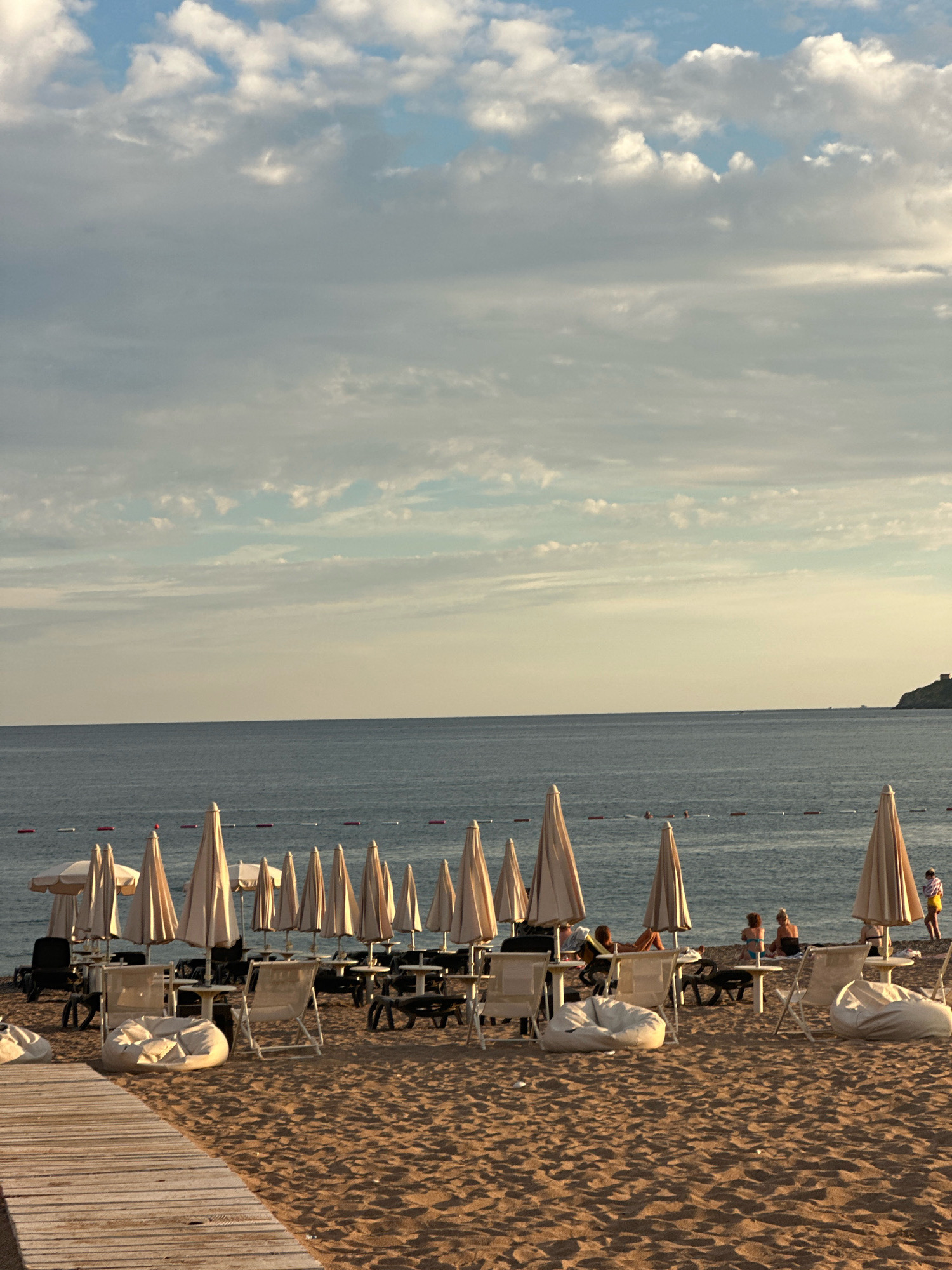 The image depicts a beach scene at what is likely the city of Boreti, Montenegro. In the foreground, a wooden walkway leads onto the sandy beach. Scattered across the sand are numerous beach chairs, tables, and large, umbrella-like sunshades. Some beanbag chairs are also visible. In the middle ground, several people are present, some sitting at tables under the sunshades, while others are relaxing on the beach. The sea stretches out to the horizon, with a subtle buoys line marking a section of the water. In the far distance, a rocky outcrop with a structure on it is visible to the right. The sky is filled with a mix of light and dark clouds, indicating a dynamic weather pattern.