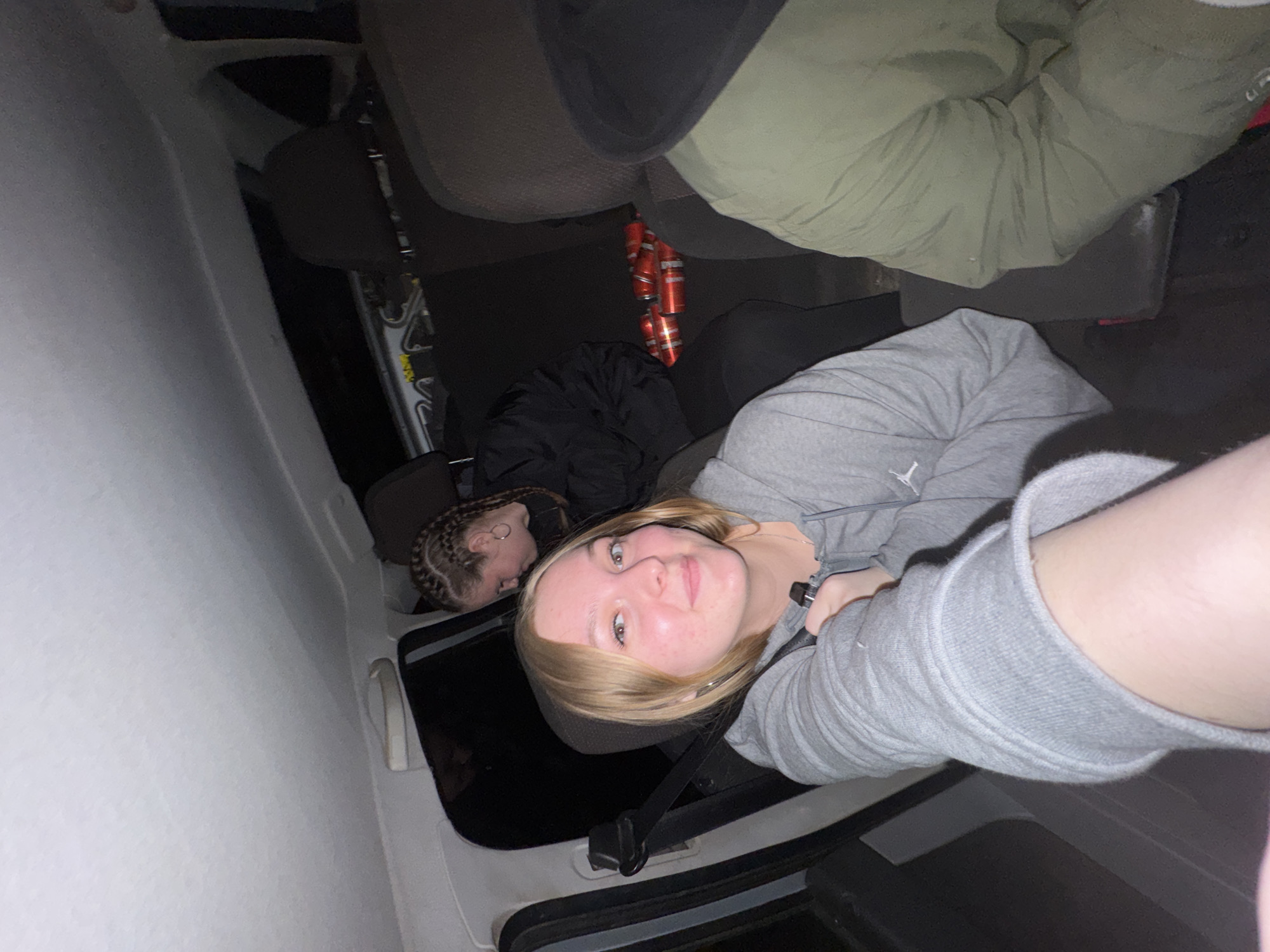 This sideways-oriented image captures two young women inside the interior of a vehicle, likely a car or van, in Chaikovsky, Russia. The perspective is a low-angle shot looking up.
In the foreground, a young woman with light blonde hair and fair skin looks directly at the camera, offering a slight smile. She has light-colored eyes and visible freckles on her face. She is wearing a gray hooded sweatshirt, and her left arm, partly clad in the same sweatshirt, is visible towards the bottom right of the frame. A seatbelt buckle is visible near her.
Behind and to the left of her, another young woman is visible. She has dark hair styled in cornrows and appears to be resting or looking down, with her head leaning against a dark surface. She is dressed in a dark jacket.
On the floor between the vehicle's seats, several red cans are visible, stacked or scattered, suggesting they are beverage cans. The vehicle's interior features dark upholstery on the seats and headrests, contrasted by lighter-colored paneling on the side. The lighting appears dim, indicating it might be nighttime or a low-light setting. The scene conveys a casual, relaxed moment, possibly during travel or a stop. No significant text is legible within the image.