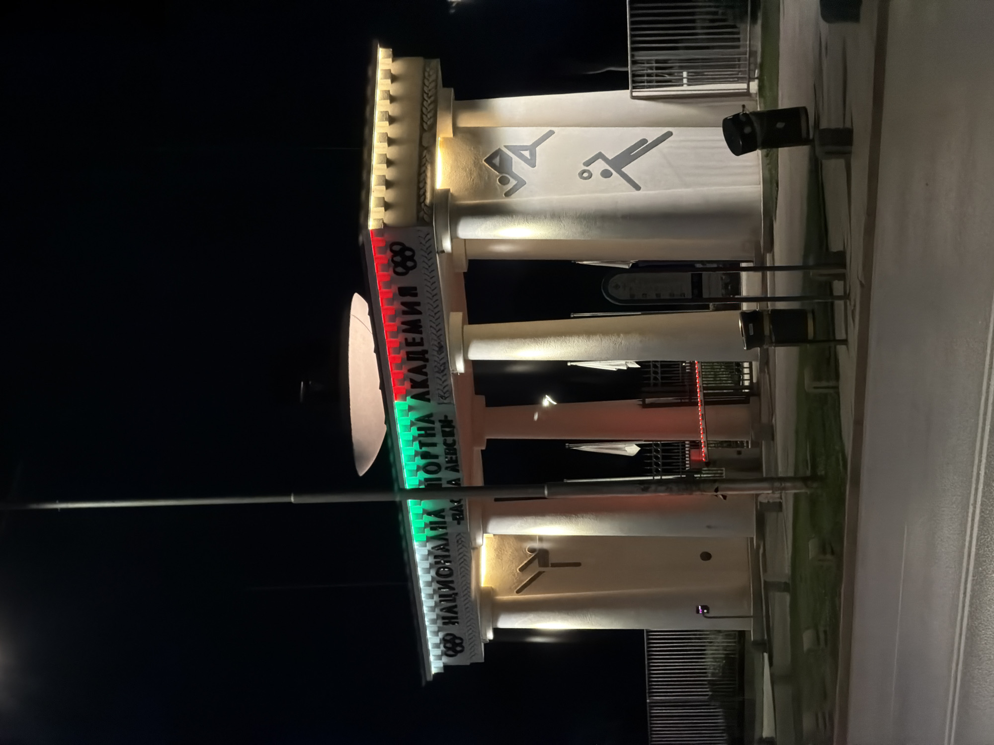 The image captures the illuminated facade of the National Sports Academy "Vasil Levski" in Sofia, Bulgaria, at night. The building features classical architectural elements including large columns and a decorative cornice, all lit by spotlights from below, highlighting their structure.
A prominent sign runs along the upper part of the building, displaying the Olympic rings emblem on the left. The sign's Cyrillic text, "НАЦИОНАЛНА СПОРТНА АКАДЕМИЯ В. ЛЕВСКИ" (National Sports Academy V. Levski), is illuminated in bright red and green segments, reflecting the colors of the Bulgarian flag.
On the building's walls between the columns, several stylized black pictograms depict athletes engaged in sports, including what appears to be a swimmer/diver and a player with a ball.
The foreground shows a paved area, likely a road or sidewalk, with white markings, and a narrow strip of green grass bordering the building. Several dark-colored bins or street furniture are visible near the base of the structure. A large street light or lamp fixture is present on the left, contributing to the bright artificial illumination that contrasts with the dark night sky. The scene depicts a significant sports institution brightly lit, suggesting its importance even after dark. No people or specific activities are visible.