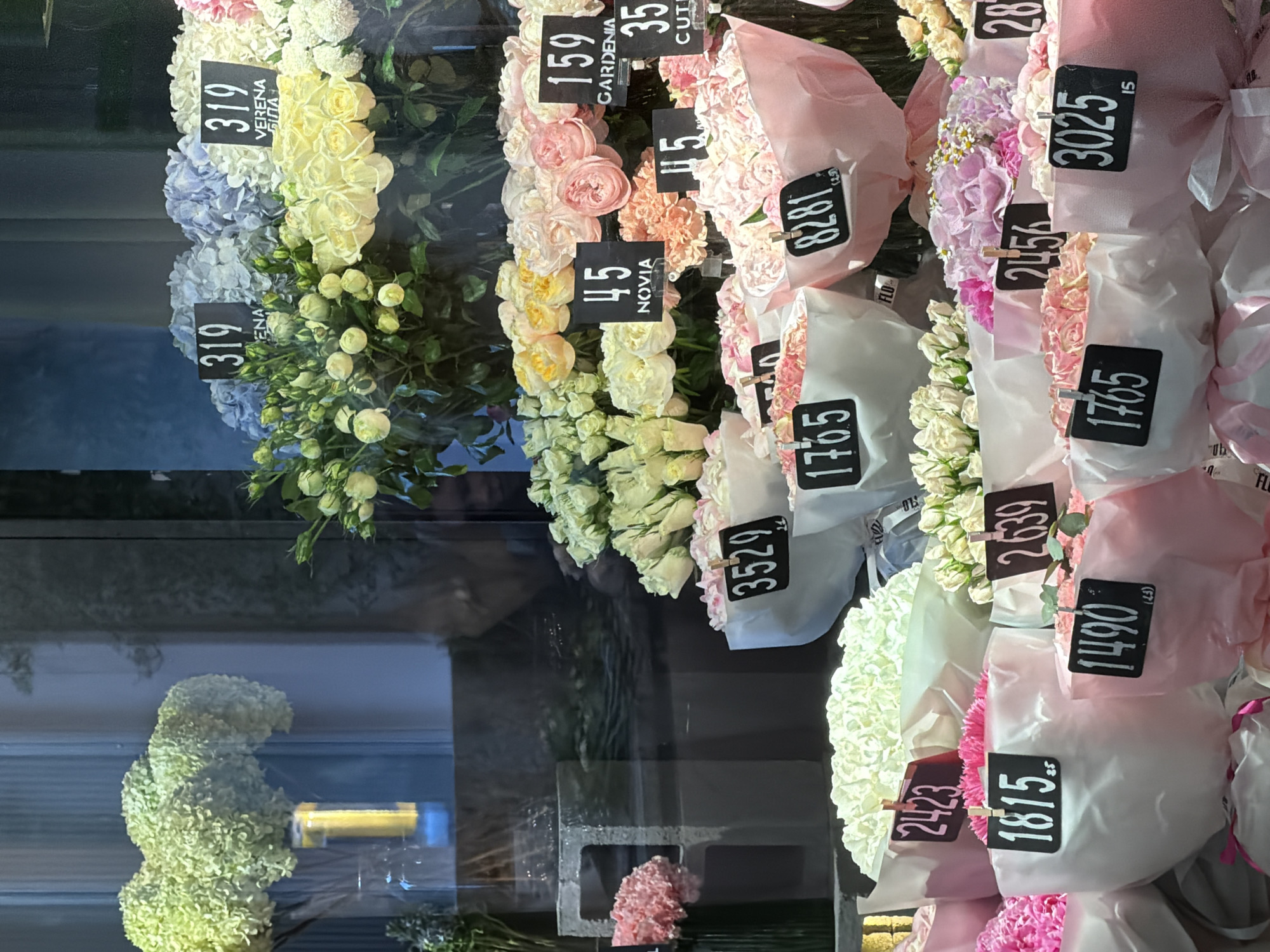 The image displays a vibrant flower shop window in Kyiv, Ukraine. The foreground is filled with an assortment of bouquets of various flowers, predominantly roses and hydrangeas, in shades of pink, white, yellow, and blue. Many bouquets are wrapped in white or pink paper and have price tags attached with clips. The price tags display numbers like "319", "159", "45", "115", "3529", "2423", "1815", "2639", "1765", "8281", "2456", "1490", "3025", and "28". Some tags also have text, such as "VERENA", "NOVIA", "GARDENIA", and "CUTI". In the background, more flowers are visible, including large bunches of light green hydrangeas in a vase with a yellow cylinder. The reflection of the interior of the shop and possibly people can be seen on the glass. The overall impression is a well-stocked and attractive flower display.