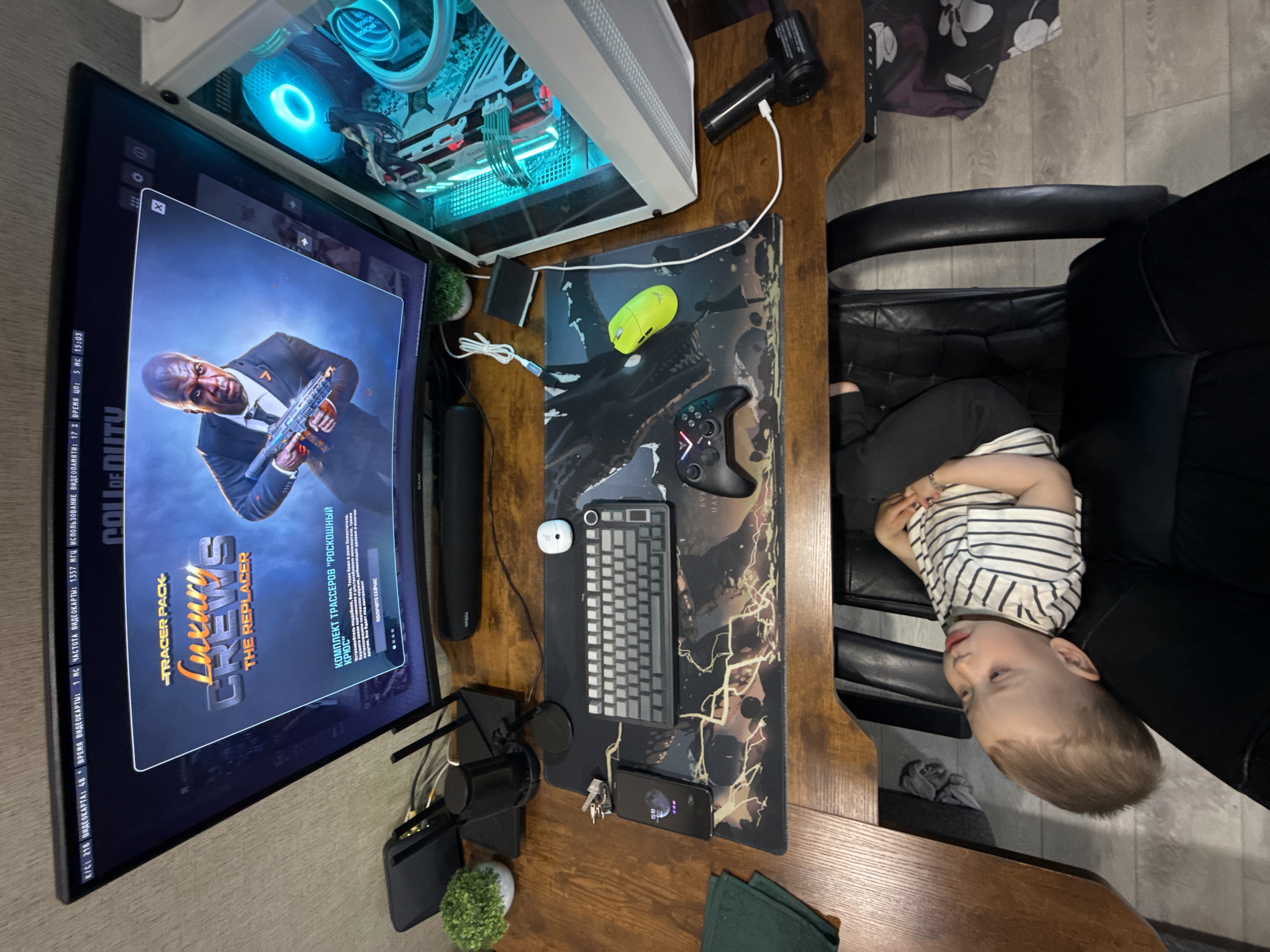 A toddler sits in a black leather chair next to a wooden desk featuring a computer setup. A large monitor displays "Call of Duty," showing an image of Terry Crews. A white PC tower with transparent side panels and blue internal lighting sits beside the monitor. On the desk, there's a black and gray mousepad with a stylized design, a black and gray compact keyboard, a white mouse, a black Xbox-style controller, a yellow wireless mouse, a smartphone, a USB microphone, and a black speaker. Small plants in pots are also present. The flooring appears to be light-colored wood or laminate. The location, Rusanovo, Russia, is inferred.