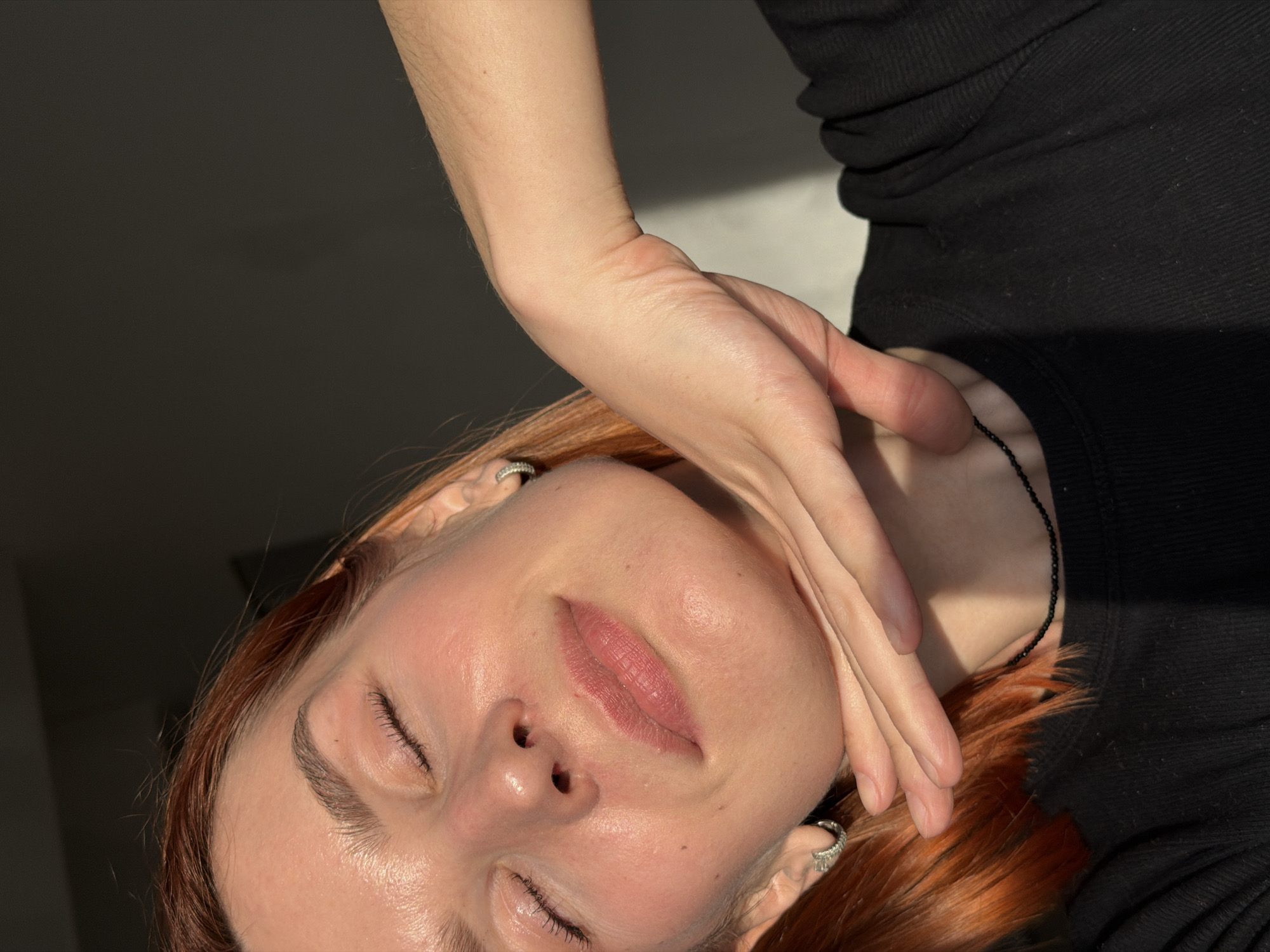 A woman with closed eyes lies on a surface, her head tilted to the left. Her skin tone is light, and she has visible eyelashes and eyebrows. Her lips are closed and have a pinkish hue. She has earrings in both ears. Her hand is placed near her neck, with her fingers slightly curved. She is wearing a black, ribbed shirt or top. A thin black necklace is visible on her neck. The background is a mix of light and dark, suggesting an indoor setting with some sunlight. The location is presumed to be Chaikovsky, Russia.