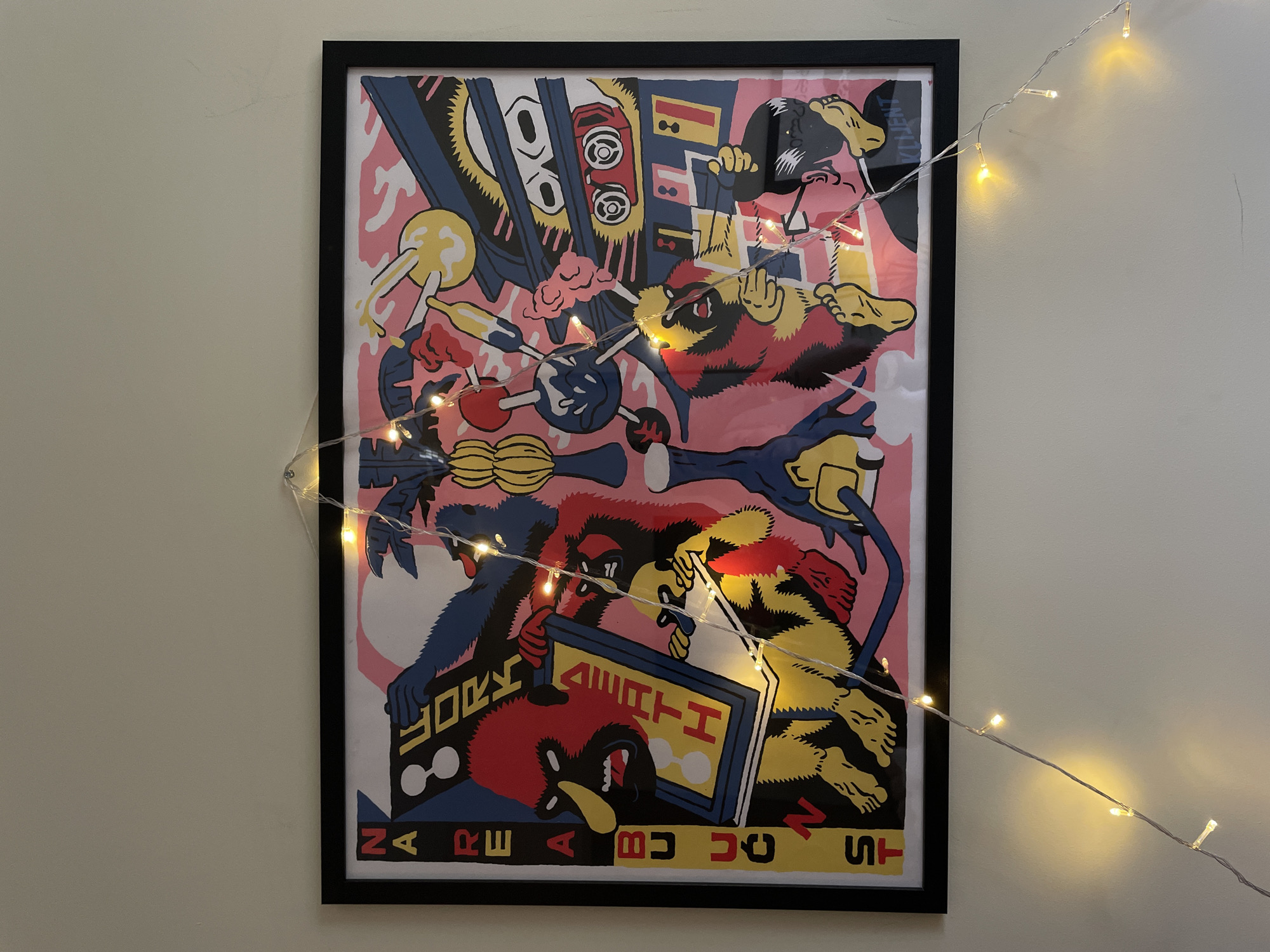 A framed, colorful illustrative poster hangs on a light beige wall. The poster depicts a chaotic scene of cartoonish monsters, with vibrant reds, blues, yellows, and pinks. The poster has a black frame. A string of fairy lights is draped around the poster, partially obscuring the artwork. The setting appears to be indoors. Based on the prompt, if this was located in the city of Vracar Urban Municipality, Serbia, this would be displayed inside someone's home, office, or some similar interior space.