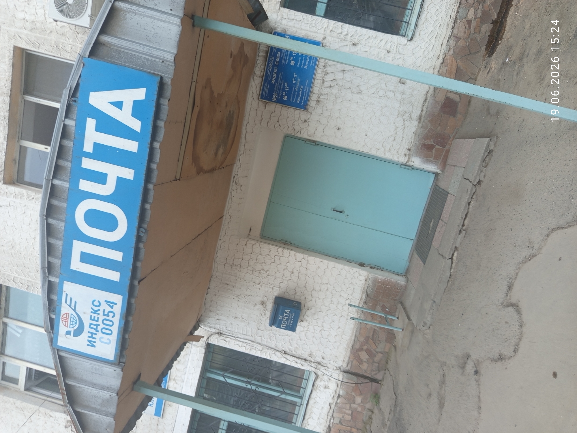 The image shows the entrance to a postal office in Bishkek, Kyrgyzstan. Above the entrance, a blue sign displays the word "ПОЧТА" (Post Office) in large white letters, along with a logo and the index number "C0054". To the left of the entrance, a blue mailbox is affixed to the textured white wall. Another blue sign to the right of the entrance provides operating hours and other information in Russian and possibly Kyrgyz. The building has a white, textured facade with a stone-like lower section. The entrance features light blue double doors. The pavement in front of the building is cracked and uneven. The overall scene suggests a functional, everyday urban environment.