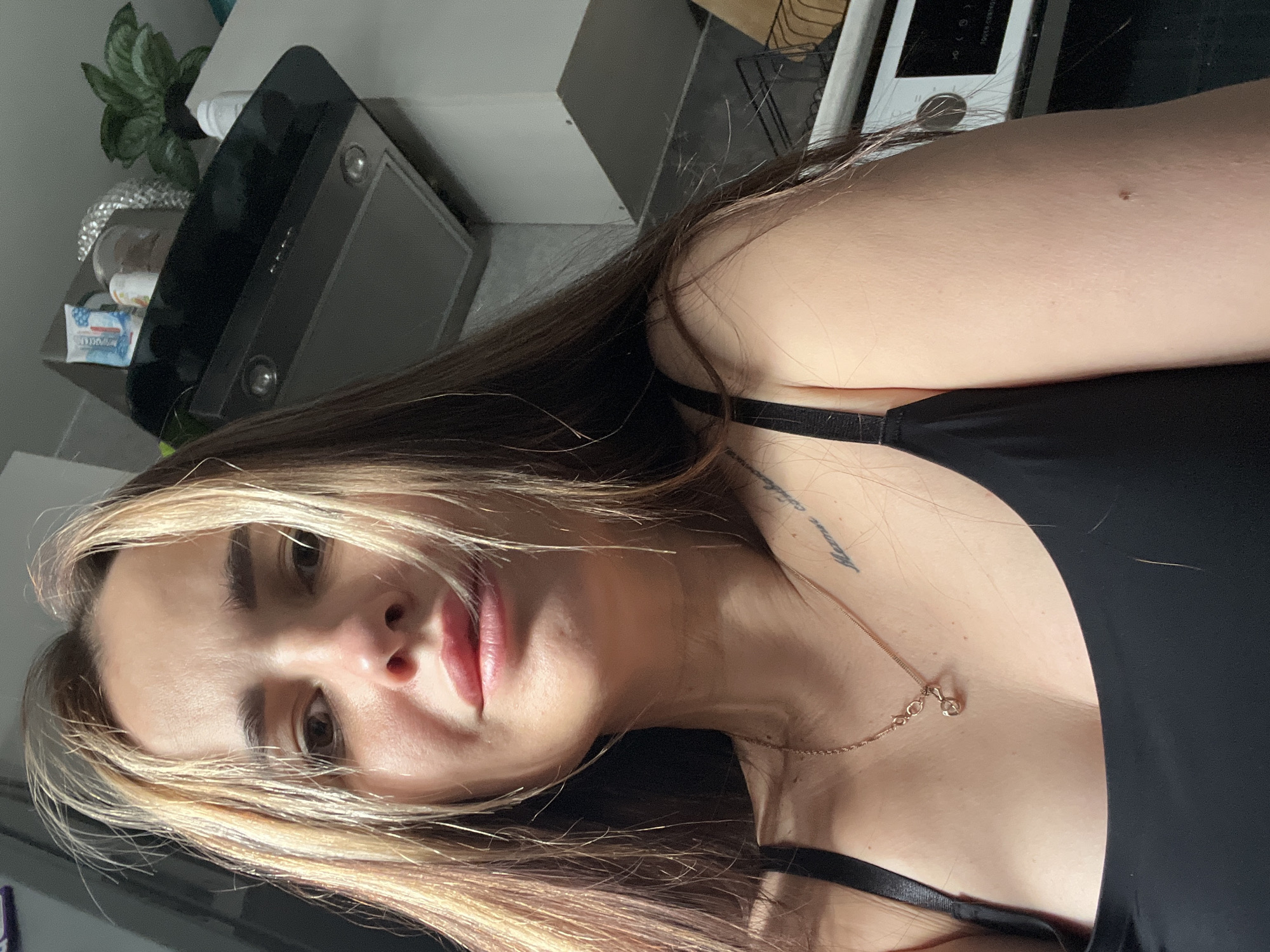 A close-up selfie of a woman with light brown and blonde hair, fair skin, and a black top. She is wearing a delicate gold necklace. A tattoo is visible on her chest near her left shoulder. The background shows a kitchen area with a black range hood, a small potted plant, and other kitchen items. The location is not specified.