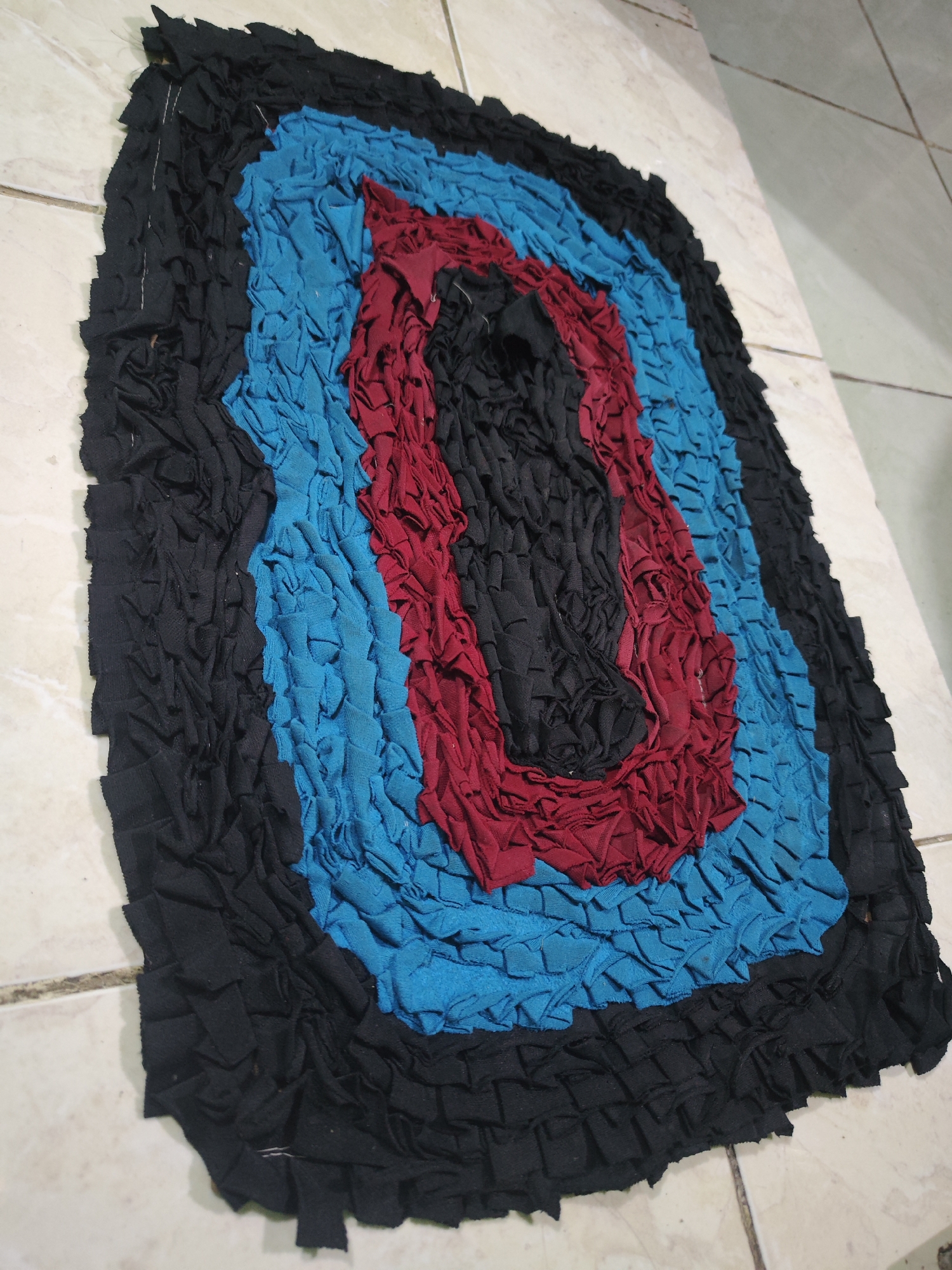 A handmade rug with concentric circles of black, blue, and red fabric strips is displayed on a tiled floor. The rug is made from cut fabric pieces that are bunched and layered to create a textured surface. The outermost layer is black, followed by a blue layer, then a red layer, and the innermost section is also black. The rug appears to be placed on a light-colored, tiled floor in what could be an indoor or outdoor setting, given the tile style. The specific location in Sroyo, Indonesia, is not visually identifiable from the image. There are no people or discernible actions taking place.