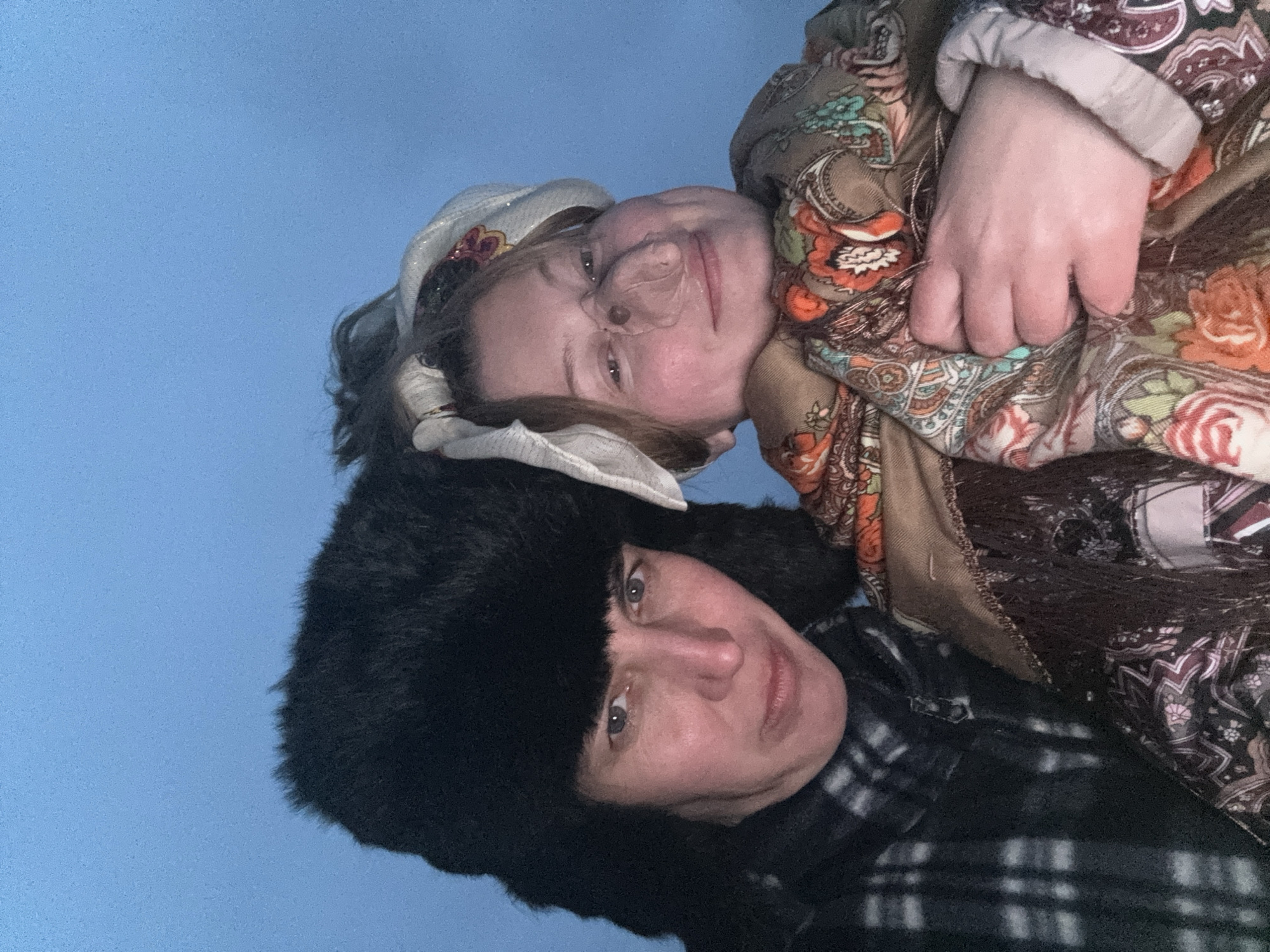 The image, rotated 90 degrees counter-clockwise, features two women posing for a close-up photograph against a plain, light blue background, suggesting an indoor setting or simple backdrop.
The woman positioned on the right side of the frame (when viewed in the original orientation) appears younger. She wears a light-colored headscarf, possibly white or cream, with subtle metallic or embroidered details, revealing dark hair. She is also dressed in a large, patterned scarf or shawl with a traditional design incorporating shades of brown, orange, green, and maroon. A portion of her patterned sleeve is visible. She has a subtle smile and light-colored eyes. Her left hand is visible, resting on her patterned clothing.
The woman positioned on the left side of the frame (when viewed in the original orientation) appears older. She is wearing a prominent black fur hat, characteristic of an ushanka style, and a dark plaid shirt or jacket. She has a neutral expression and looks directly at the camera with light-colored eyes.
Both women exhibit fair complexions and are dressed in warm attire, which aligns with the provided location context of Вассята, Russia. There is no visible text in the image.