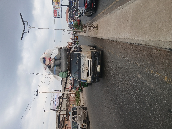 The image depicts a busy, overcast daytime scene on what appears to be an urban or semi-urban street. The perspective is from the side of the road, looking forward along the direction of traffic.
In the foreground and center, a white or light-colored utility van or small truck is heavily overloaded with goods on its roof rack. The vehicle itself appears weathered, with visible dirt or discolored patches on its front and sides. Its front grille and headlights are visible, and the windshield reflects the sky. The load on top consists of numerous large sacks or bags, predominantly white and grey/blue, piled high and seemingly secured with ropes or tarps. The load significantly exceeds the height of the vehicle's cabin.
The vehicle is traveling on a two-lane asphalt road. Yellow dashed lines are faintly visible on the road surface, indicating lane divisions. To the right of the road, a raised concrete curb separates the main thoroughfare from a narrow concrete sidewalk or median, which shows some minor debris or sparse vegetation.
Along the left side of the road, a variety of buildings, possibly shops or residential structures, line the street in the mid-ground and background. Multiple utility poles with a dense network of power lines stretch across the sky. Two prominent billboards are visible:
1. In the mid-ground, a large billboard displays an advertisement featuring the brand name "LAZIZ" in large red letters, with "QUALITY" and "TASTE" written below it. The billboard also shows images of food products, appearing to be fried chicken pieces, french fries, and soft drinks.
2. Closer to the foreground, another billboard stands with what appears to be a graphic and some text, which is largely unreadable but includes script that resembles Arabic or a similar language.
Other vehicles are visible on the road: a dark-colored car is partially seen directly behind the overloaded truck, and another light-colored van or small bus is parked or moving further down the road on the left side. Traffic appears moderate.
The sky is uniformly overcast with grey clouds, indicating a cloudy day, but the scene is well-lit, characteristic of daytime. Modern streetlights with twin rectangular fixtures are mounted on tall poles along the roadside.
Visible text includes:
* On the billboard: "LAZIZ", "QUALITY", "TASTE".
* On the other billboard: Some unreadable text, possibly in Arabic script, and "FRESH".
* On the overloaded truck's side: Partially visible text, possibly "EXPRESS" or similar.
* On the license plate of the dark car behind the truck: "LAGOS" is partially visible.