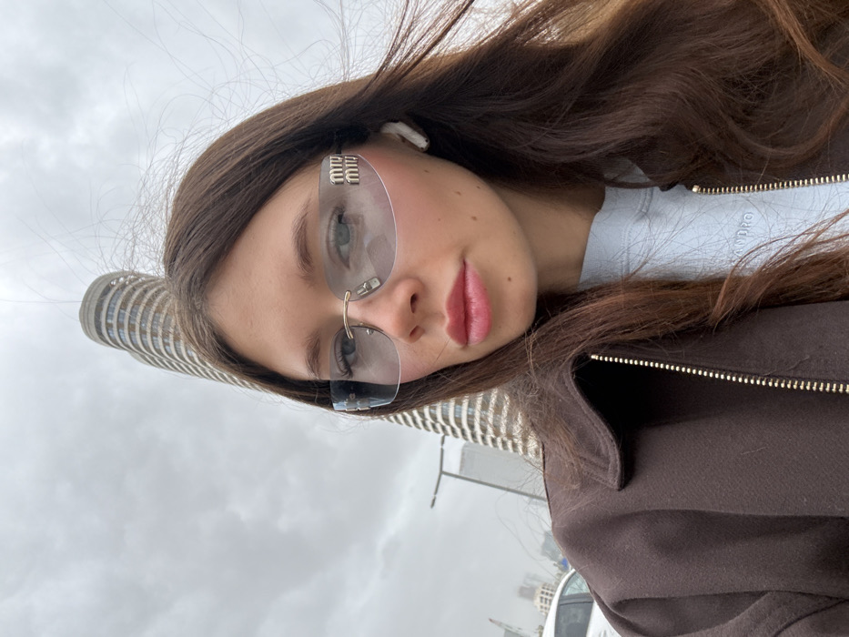 The image displays a close-up, selfie-style portrait of a female individual outdoors. She has long, dark brown hair and is looking directly at the camera. She wears light blue-tinted, rimless Miu Miu sunglasses with gold-colored frames and a white wireless earbud in her right ear. Her lips are made up with pink lipstick. She is dressed in a dark brown collared jacket with a visible gold zipper, over a light blue crew-neck sweatshirt. In the background, prominently featured is the Levinstein Tower, a tall, cylindrical high-rise building with distinctive curved balconies, locating the scene at 23 Menahem Begin Road in Tel Aviv-Yafo, Israel. The sky is overcast and grey. A portion of a white vehicle and a street light structure are visible on the left side of the frame.