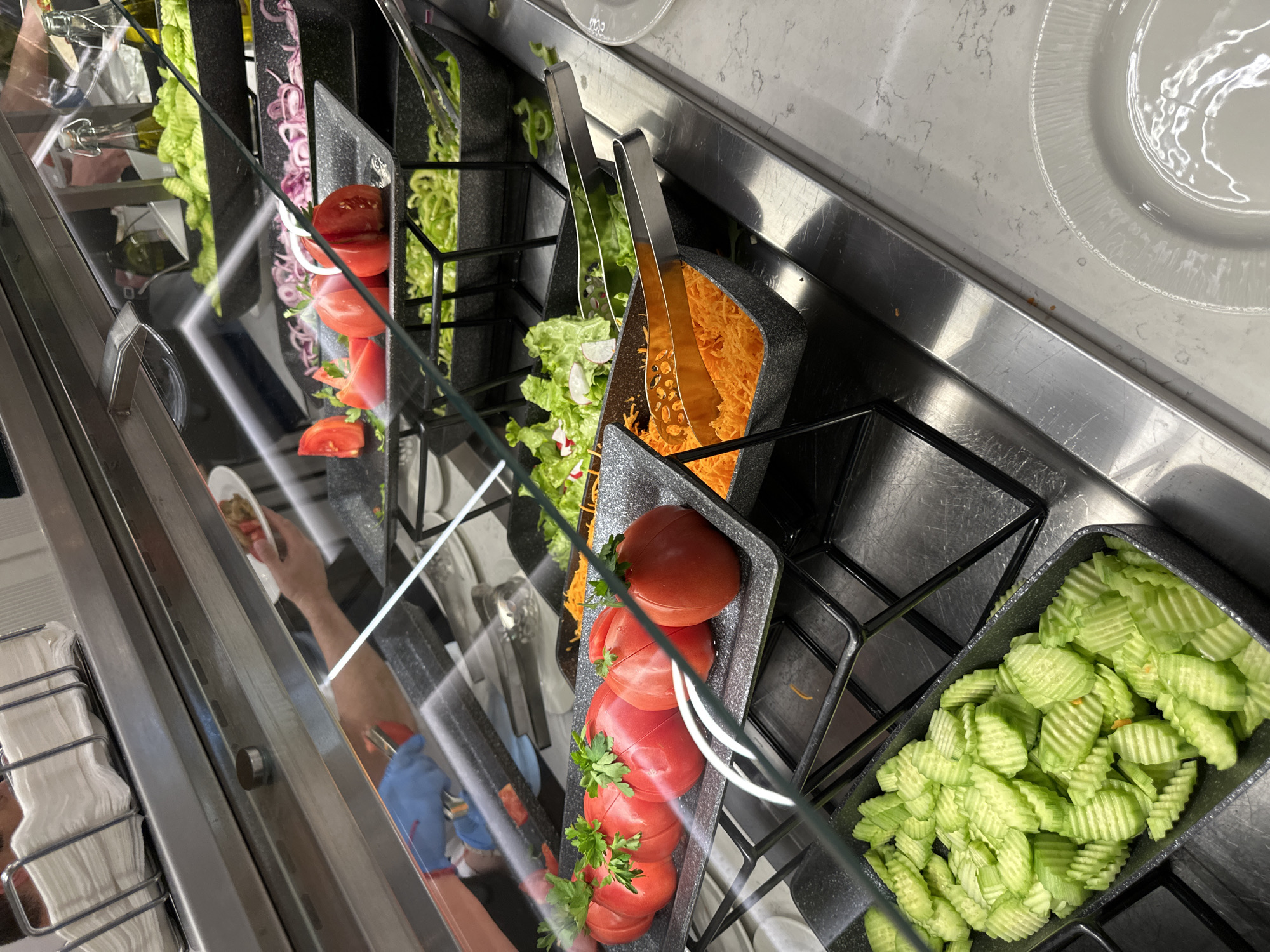 A salad bar is stocked with various ingredients in a restaurant or cafe, likely in Kazanlak, Bulgaria, based on image origin. The camera angle is from above, looking down into the salad bar through a protective glass shield.
From left to right, ingredients are presented in long, rectangular, dark gray containers held by a black metal frame. They include:
* Lettuce varieties
* Thinly sliced red onions
* Tomatoes cut into wedges, garnished with sprigs of parsley
* Shredded carrots
* Sliced cucumbers with a ridged texture.
Tongs are inserted into the containers of lettuce and shredded carrots. The salad bar setup is stainless steel. Napkins are in a metal holder to the left of the salad ingredients. A reflection shows a person holding a plate with food, with another person in a blue glove appearing to be refilling the salad bar.