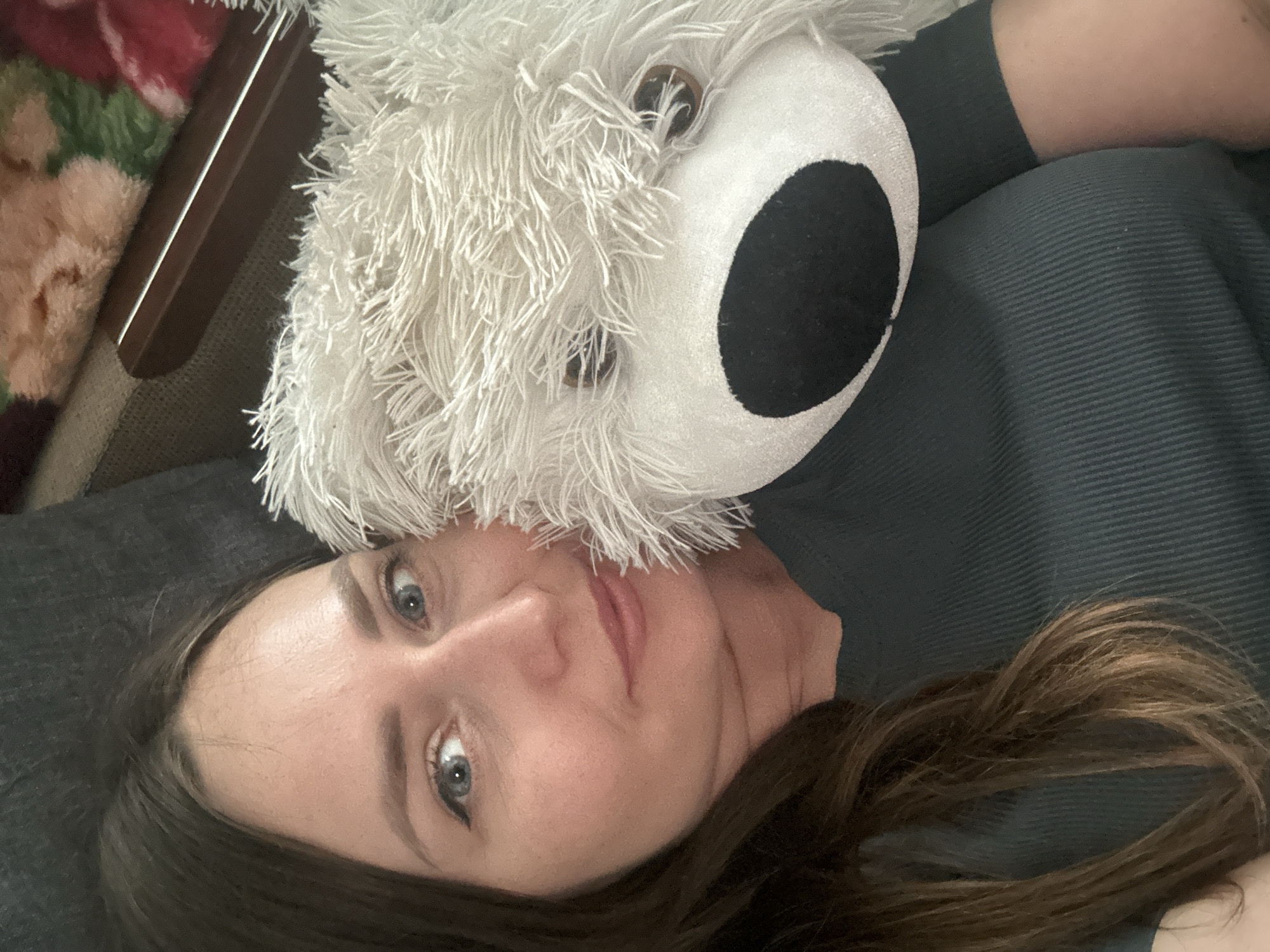 A woman with long, dark brown hair is lying down, holding a fluffy white teddy bear close to her face. She has blue eyes and is looking towards the camera. She is wearing a dark green ribbed t-shirt. The teddy bear has a black nose and brown button eyes. The background appears to be a couch with a textured, dark fabric and a blanket with floral patterns in pink, green, and red. The image does not provide any information about the city of Bila Tserkva, Ukraine.