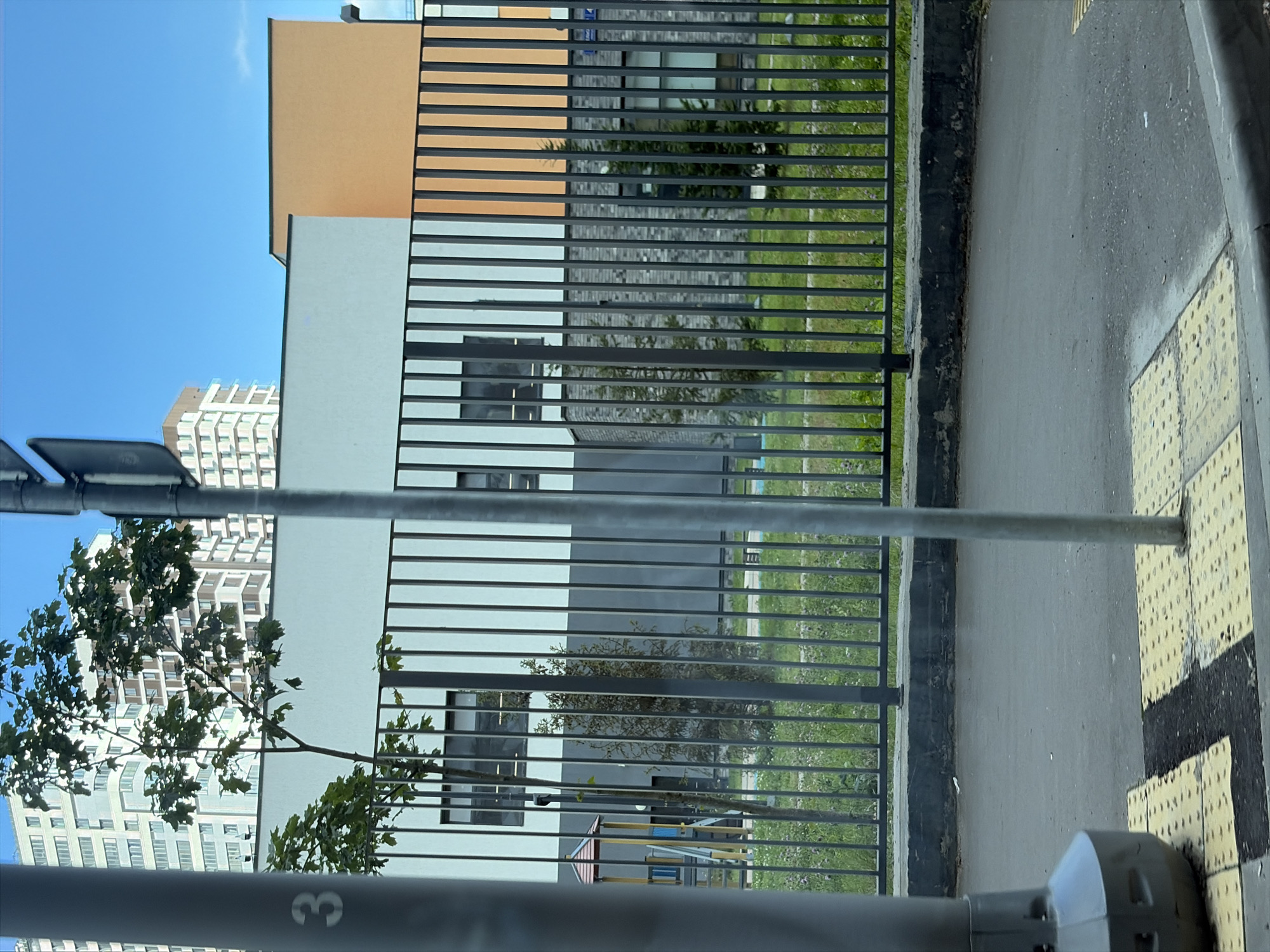 The image depicts a street-level view of a modern urban area, likely in Moscow, Russia, based on architectural style and the presence of Cyrillic lettering on a sign pole. In the foreground, a grey sidewalk with yellow tactile paving marks is visible, along with a section of asphalt road. Two metal poles, one thick and grey on the left and a thinner silver one in the center, stand prominently. A black metal fence runs horizontally across the mid-ground, separating the sidewalk area from a grassy patch and a building complex.
Behind the fence, a low, grey concrete border is visible, followed by a strip of green grass with scattered wildflowers. The building complex features a modern architectural design with contrasting facade colors. The left section of the building is light grey with multiple rows of windows. Adjacent to it is a section with an orange facade. Further to the right, a darker, textured facade, possibly brick or stone, is visible with more windows. A small playground structure with a red and yellow roof is partially visible to the left of the building, behind the fence.
In the background, a tall, multi-story residential building with a light-colored facade and numerous windows dominates the upper left portion of the image. A clear blue sky with a few faint clouds is visible above the buildings. A partial view of a street sign pole with Cyrillic characters, possibly indicating street names or directions, is present in the upper left quadrant. The overall scene suggests a newly developed residential or educational area with modern infrastructure. No people are visible in the image.