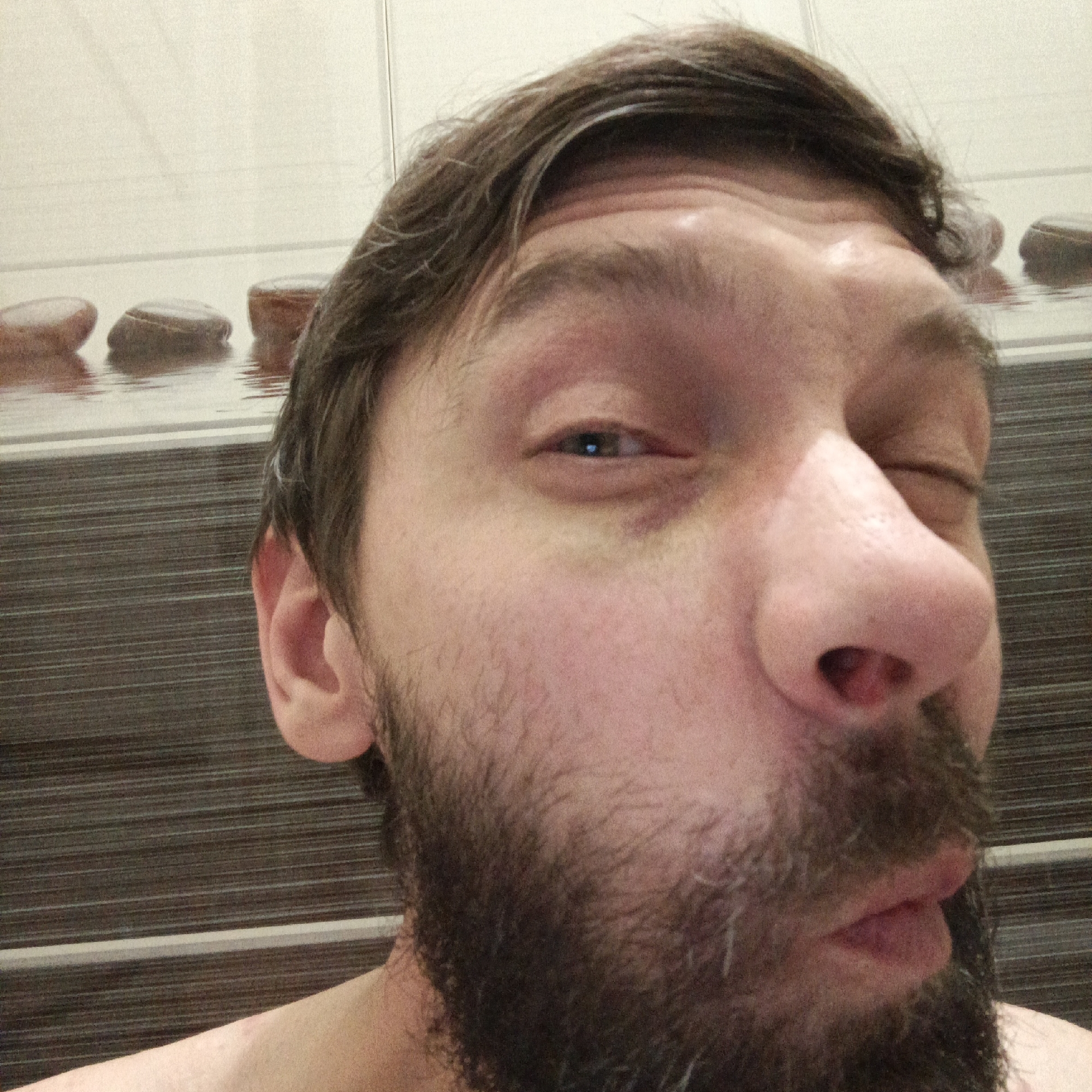 A close-up selfie captures a man with light skin, a dark beard, and brown hair. He is squinting one eye, puckering his lips, and looking directly at the camera. The background appears to be a bathroom wall with a horizontal striped pattern and a row of decorative stones near the top. There is no evidence that the image was taken in Марковский, Russia.