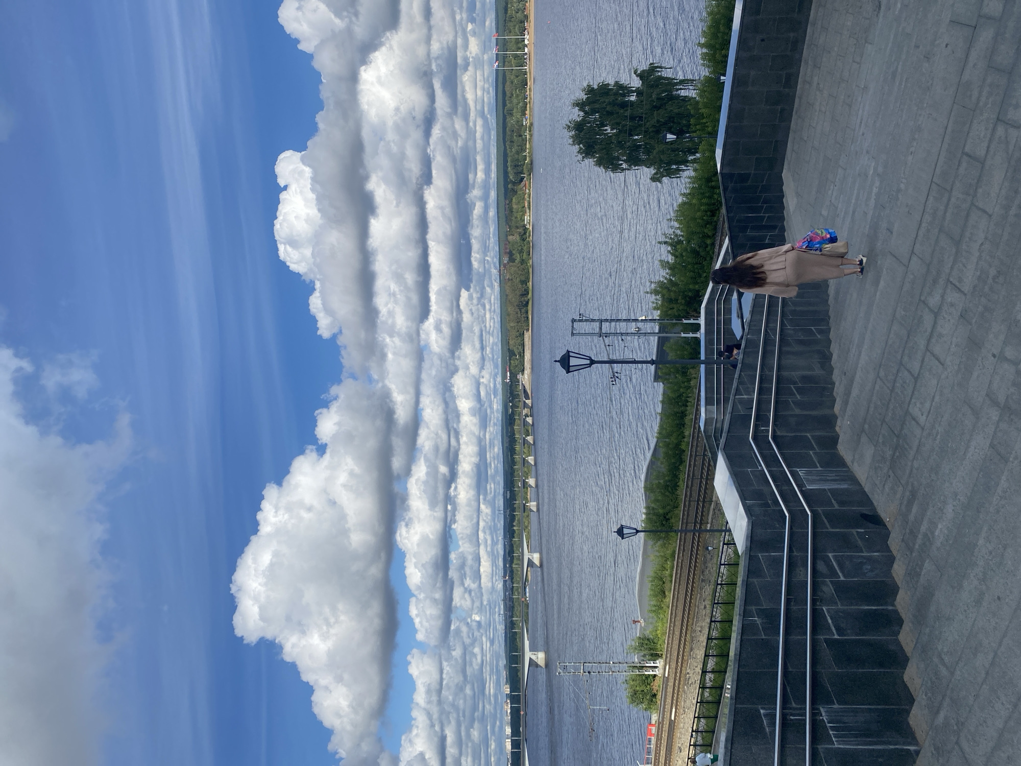 A woman walks up a wide outdoor staircase towards a river and a long bridge. The staircase is made of gray stone with dark stone walls and metal railings. The woman is wearing a beige knit outfit and carrying a tote bag with colorful shopping bags. She is walking away from the camera, towards the river.
The river is wide and calm, with ripples on its surface. A long, multi-span bridge crosses the river in the distance, connecting the two shores. The far shore is lined with trees and buildings, indicating an urban area. The sky is blue with large, fluffy white clouds.
In the middle ground, between the staircase and the river, there are railroad tracks, a few streetlights, and some scattered trees. There is a single, large green tree to the right of the frame, near the riverbank. The overall scene appears to be a scenic viewpoint overlooking the Kama River in Perm, Russia, possibly near the Perm-1 railway station or the river port area, given the presence of the tracks and the view of the bridge. The lighting suggests it is daytime, possibly late afternoon given the slightly muted tones.