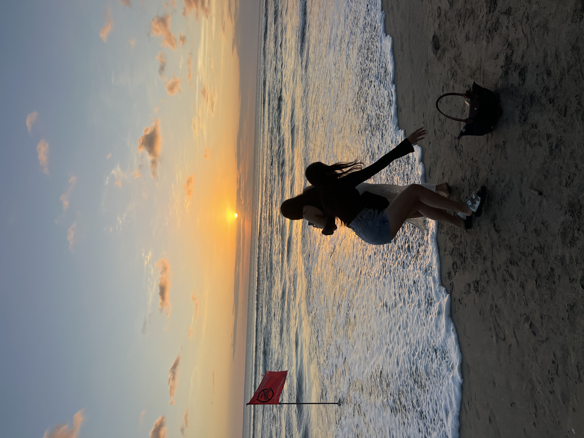 Two women are standing on a sandy beach in Kuta, Indonesia, at sunset. The sky is a gradient of orange, yellow, and blue with scattered clouds. The sun is a bright orb on the horizon, reflecting on the ocean waves. A red flag with a symbol on it is planted in the shallow water to the left of the frame. The woman on the left is embracing the woman on the right, who is reaching out towards the water. A dark-colored bag is placed on the sand near the woman on the right. The sand is dark and appears wet near the water's edge, with footprints visible. The ocean has gentle waves breaking onto the shore.