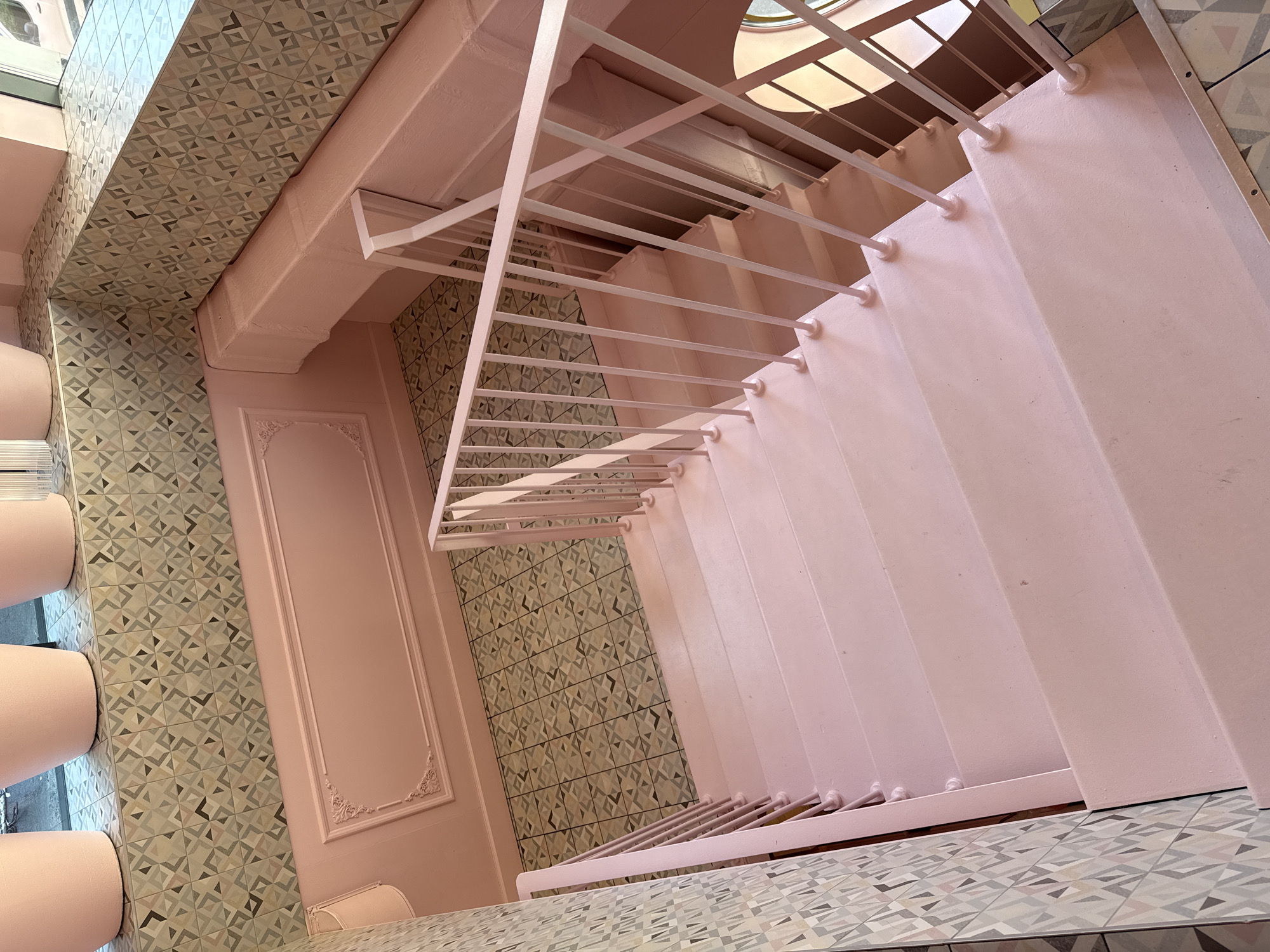 The image shows a bright pink staircase with a white railing, descending from what appears to be an upper floor. The staircase is constructed of metal with a smooth, painted finish. The steps are wide and the risers are low, suggesting a comfortable ascent or descent. The railing is made of thin metal posts and a handrail, all painted white.
The walls adjacent to the staircase are also painted pink. To the left of the staircase, there is a decorative wall panel with ornate molding. The floor at the bottom of the stairs is tiled with a geometric pattern in shades of grey, beige, and muted green. Above the pink wall, there is a band of the same geometric tile pattern.
The overall impression is of a clean, modern, and aesthetically designed interior space. There are no people or animals visible in the image. The lighting suggests an indoor setting, possibly with natural light coming from a window or opening not fully visible in the frame. The building appears to be relatively new or recently renovated, given the pristine condition of the staircase and walls.