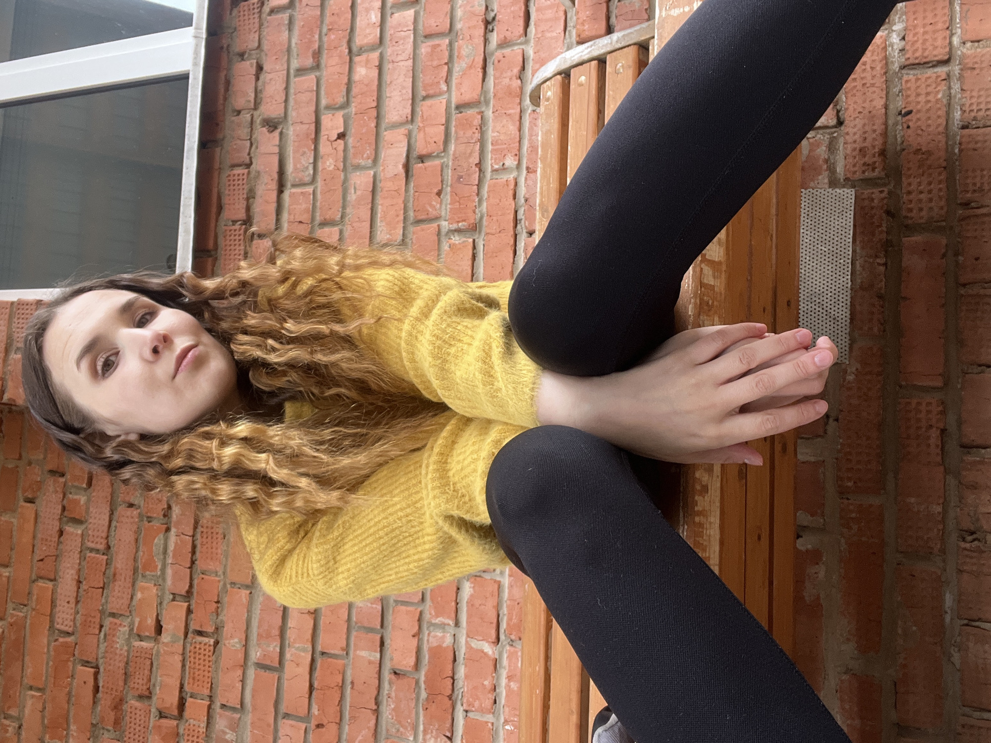 A young woman with light skin, brown hair, and a headband is sitting on a bench in front of a red brick wall, possibly exterior wall to a building. She is wearing a yellow sweater and black leggings. Her legs are positioned over the back of the bench, and her hands are clasped together near her feet. A window is visible in the upper left corner of the frame. This location is Votkinsk, Russia.