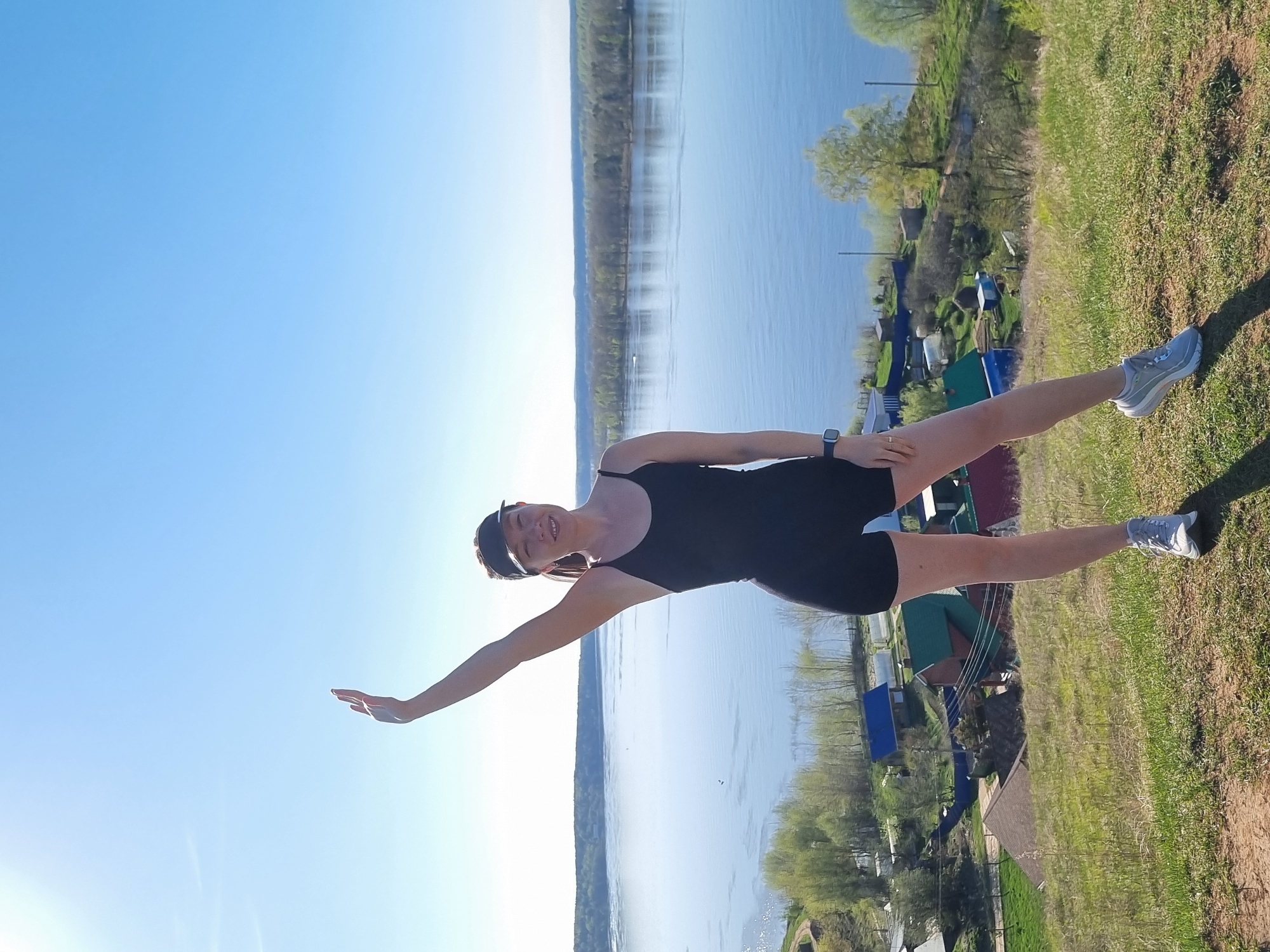 A woman is standing on a grassy hill overlooking a body of water, possibly in Ольховка, Russia. She is wearing a black bodysuit, a baseball cap, and sneakers. She has her left arm extended out to the side. The water is calm and reflects the sky. There are trees and buildings on the opposite side of the water.