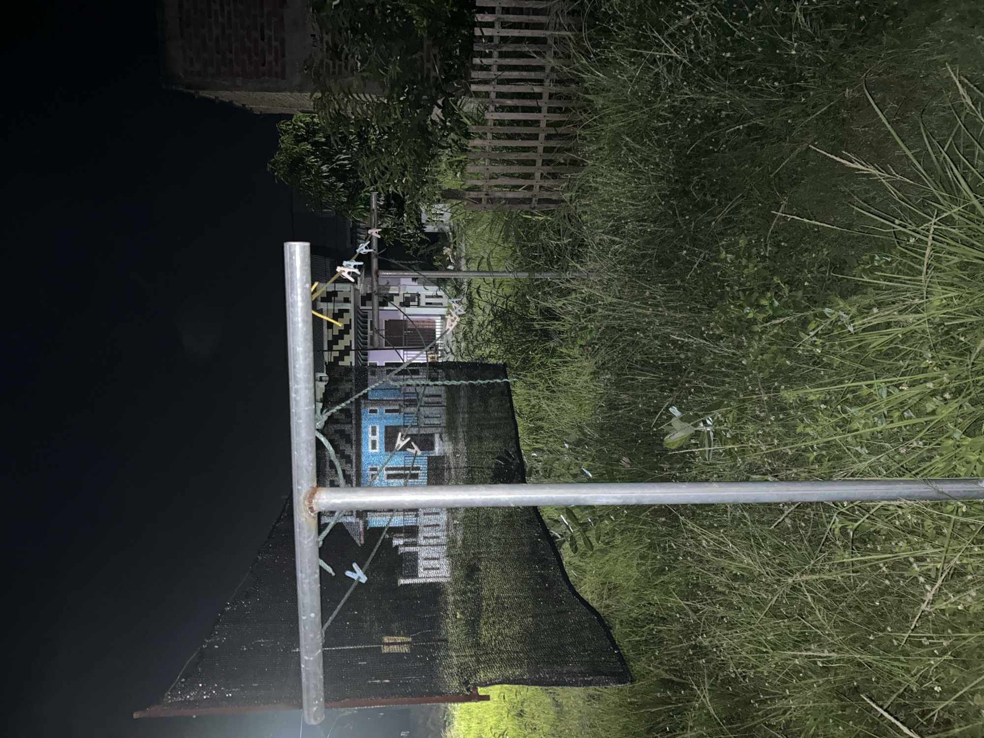 The image depicts an outdoor scene in Lhokseumawe, Indonesia, likely at night or in low light conditions. Tall grass is prominent in the foreground and background. A metal clothesline is visible in the center, with a dark mesh fabric hanging from it. Clothespins are attached to the clothesline. A partially visible house is behind the clothesline. The house appears to be blue and white. In the distance, a wooden fence and trees can be seen. The upper-left of the image is dark, with the suggestion of a building in shadow.