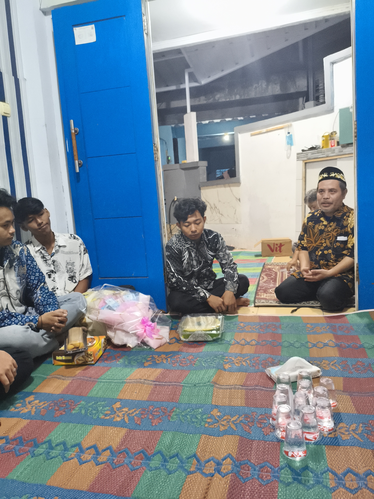 In Depok, Indonesia, a group of four young men and one older man are gathered indoors, seated on a colorful woven mat. The room appears to be a living area, with a large blue double door dominating the left side of the frame. The older man, wearing a traditional peci hat and a patterned shirt, is seated on a rug and appears to be speaking. The younger men are seated on the mat, with two on the left side of the frame and one in the center. A bouquet of pink and white flowers, wrapped in plastic, sits in front of the two younger men on the left. A box of snacks is also visible near them. In the foreground, a cluster of plastic cups, some filled with a clear liquid, is arranged on the mat. The background shows a partially open room with exposed ceiling beams and lighting, suggesting a simple interior setting.