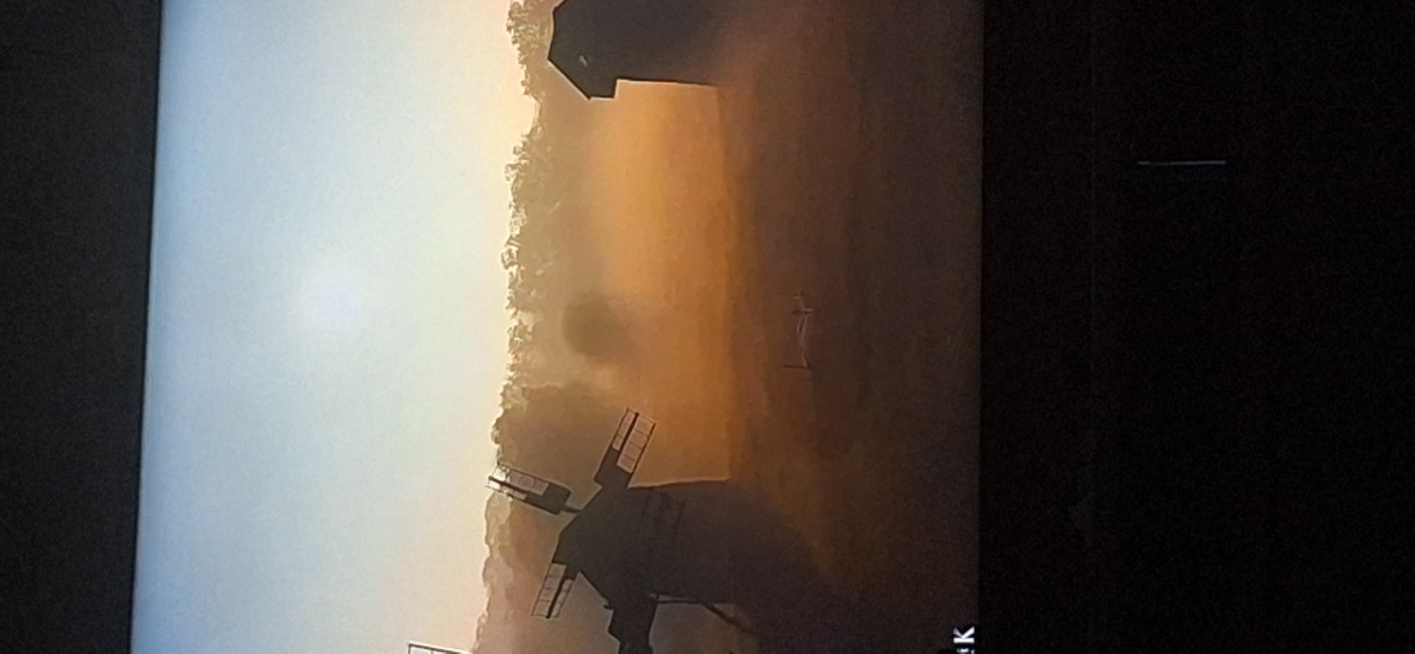 The image depicts a sunrise over a misty landscape with two windmills and a small building. The sky is a gradient of light blue and orange. In the foreground, on the left, is a silhouetted windmill with four sails. To the right, further in the background, is a smaller, dark building. In the middle ground, a faint, circular object is visible through the mist, possibly a tree or a distant structure. A small, light-colored object, possibly a drone, is lying on the ground in the center of the frame. The bottom left corner of the image shows the letters "K". There are no people visible in the image. The scene appears to be a rural or semi-rural setting.
