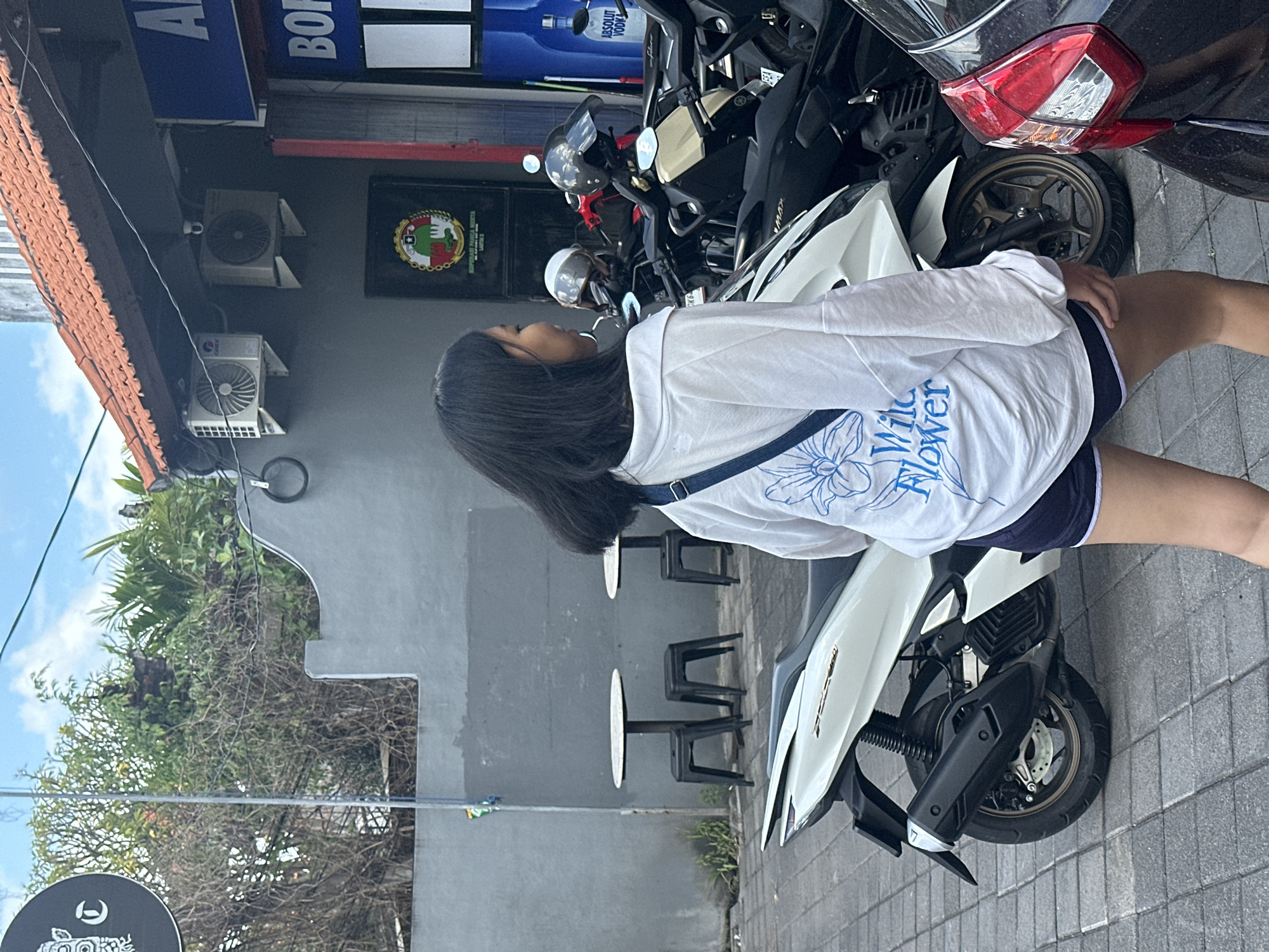 An over-the-shoulder shot captures a young Asian woman from the waist up, walking away from the camera on a paved sidewalk. She has dark hair and is wearing a white long-sleeved t-shirt with a blue floral graphic and the words "Wild Flower" printed on the back, and dark shorts. A dark blue strap crosses her chest. To her left, a white scooter is parked. Behind the scooter, several other parked motorcycles and a black car are visible. The background features a gray building with red-tiled roofing and outdoor air conditioning units. A sign with blue lettering and a bottle of Absolut Vodka are also visible on the building. Trees and vegetation are present behind the building. The scene appears to be an outdoor commercial area in North Kuta, Indonesia, given the architecture and parked vehicles.