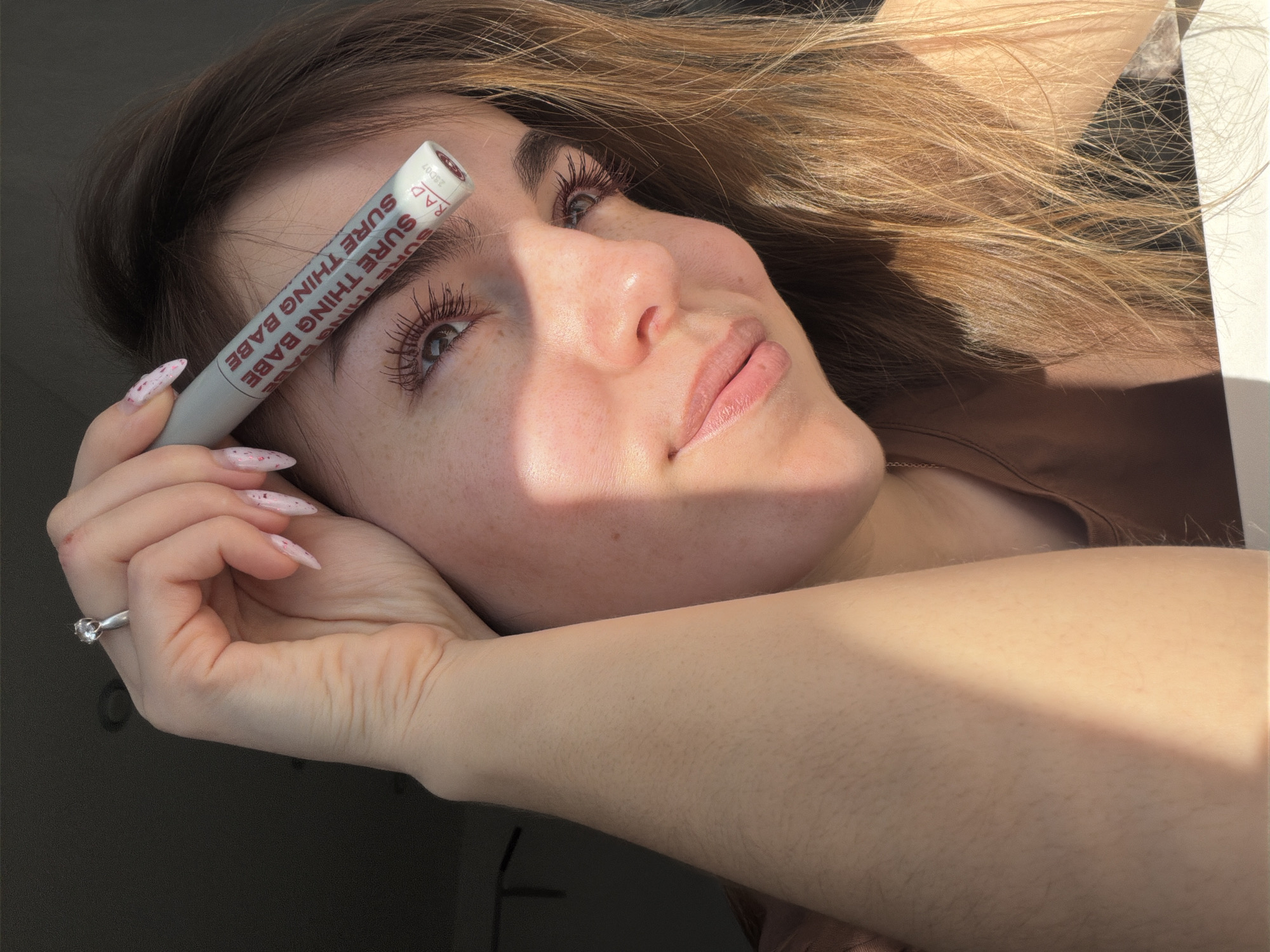 A young woman with fair skin and light brown hair is photographed in a brightly lit environment. She holds a white tube labeled "SURE THING BABE" near her forehead, between her eyebrow and hairline. Her eyes are light colored, and her long eyelashes are enhanced with a dark mascara. She has freckles on her cheeks and nose. Her lips are naturally pink and slightly glossy.
She wears a silver ring with a clear stone on her left ring finger. Her fingernails are long and painted with a light pink color and darker pink speckles. A light brown shirt is visible at her shoulder.
The angle suggests she is looking up at the camera. The background is not clearly visible but suggests an interior space. The location in Sarapul, Russia, is not apparent from the image.