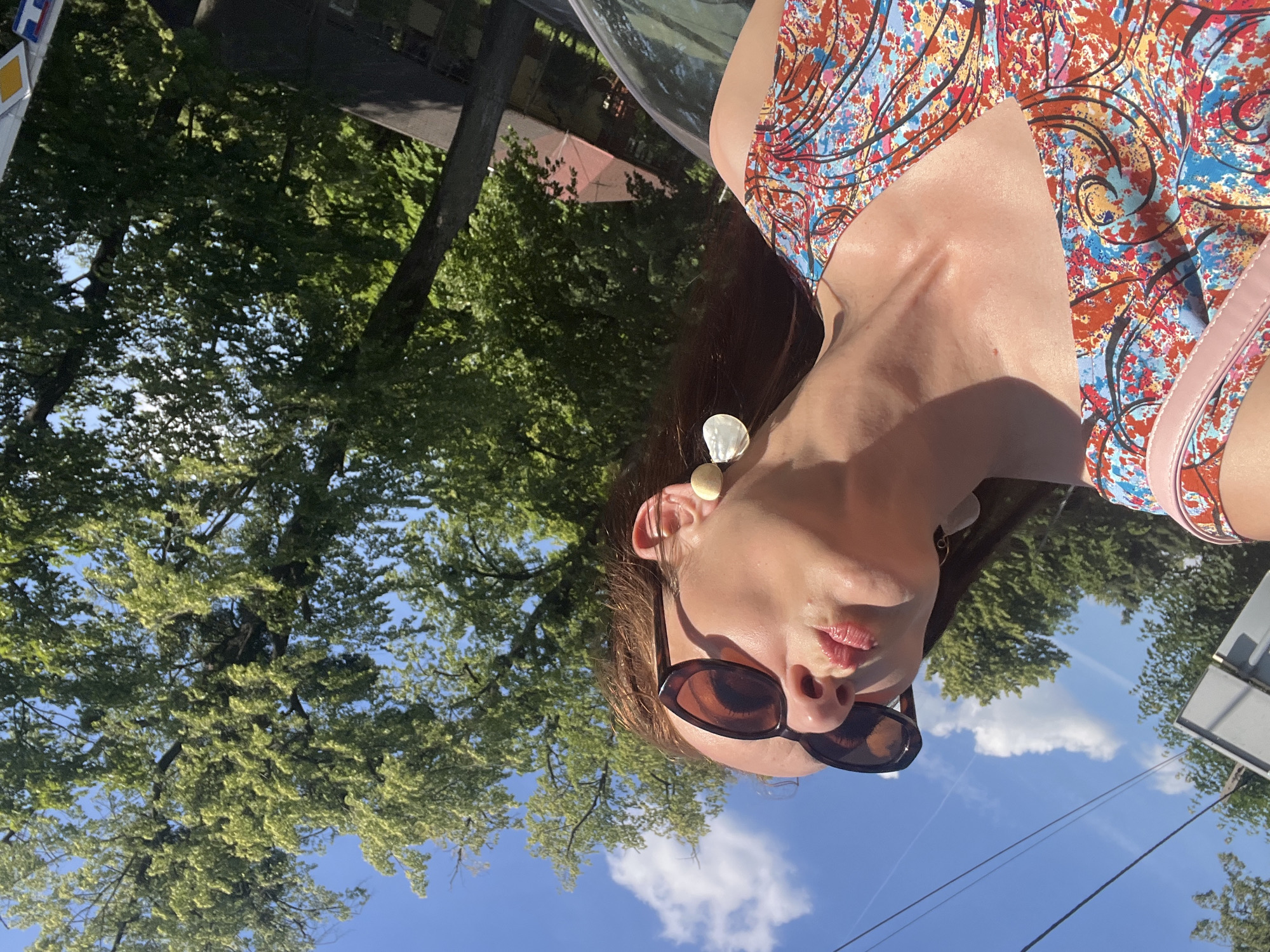 A woman wearing sunglasses and a colorful dress is captured from the shoulders up, with her head tilted back and eyes closed. She has red hair and is wearing large, shell-like earrings. The background is a bright blue sky with fluffy white clouds and lush green trees. The image appears to be taken outdoors on a sunny day, possibly from inside a vehicle due to the angle and slight reflection on the right side. The setting is not clearly identifiable as a specific city, but the presence of trees and a sky suggests an outdoor, possibly urban or suburban, environment.