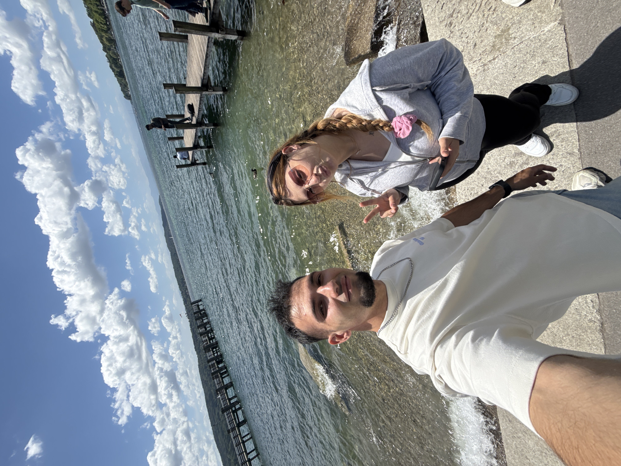 A high-angle, wide selfie shows a young man in the foreground, wearing a cream-colored t-shirt and a silver chain, with a beard and short, dark hair. To his right is a young woman wearing large pink sunglasses, a grey zip-up hoodie over a white top, black pants, and white sneakers. She is holding a black smartphone and making a peace sign with her right hand. They are standing on a concrete or stone embankment next to the clear, blue water of Starnberger See, Germany. In the background, there is a wooden pier with several people on it, and a distant shoreline with lush green trees and hills under a bright blue sky with scattered white clouds. Several ducks are visible in the water near the embankment.