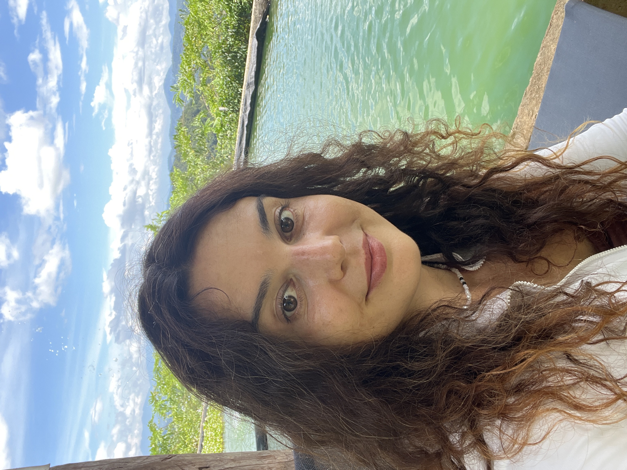 A woman with long, dark, curly hair and brown eyes is captured in a selfie taken outdoors. She is wearing a white top and a beaded necklace. She is positioned in the foreground, with a turquoise swimming pool to her right and lush green vegetation and hills in the background. The sky above is blue with scattered white clouds. The scene appears to be in a tropical or subtropical location, possibly Sơn Điền, Vietnam, given the presence of dense greenery and hilly terrain. The image has a natural, bright lighting, suggesting it was taken during the daytime.