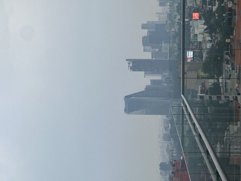 A high-angle, wide-shot photograph depicts a hazy urban landscape of Mexico City, Mexico, from an elevated vantage point. In the immediate foreground, a glass and metal railing runs diagonally from the lower left to the center, then horizontally to the right, partially obscuring the lower midground. The sky occupies the upper half of the image, appearing uniformly light blue-gray with significant atmospheric haze; a faint, diffuse circular light source is visible in the upper right.
The midground features a dense array of low-to-mid-rise urban buildings, interspersed with some vegetation. On a rooftop in the center-right midground, a digital billboard displays the text "Konami" and "535 x 100". Further to the right, another illuminated red-orange sign is visible on a distant building.
The background is dominated by a cluster of prominent skyscrapers, which are partially obscured by the haze. Key identifiable structures include Torre Reforma, a distinctive triangular skyscraper, positioned on the left side of this cluster, and Torre BBVA México, identifiable by its "BBVA" logo near its summit, situated to the right of Torre Reforma. Other less distinct high-rise buildings extend the skyline into the distance. No individuals are discernible within the frame.