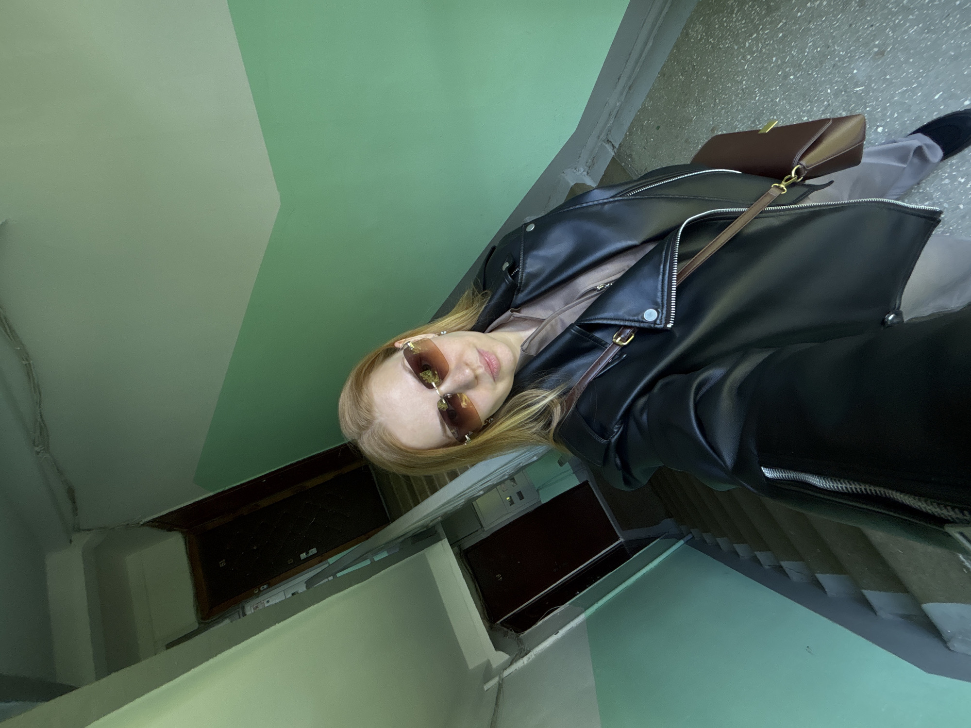 A woman wearing sunglasses and a black leather jacket stands in a stairwell. She is also wearing a beige top and carrying a brown purse. The stairwell has green walls and concrete stairs. The photo appears to be taken from a low angle. It is unknown whether the location is in Rusanovo, Russia.