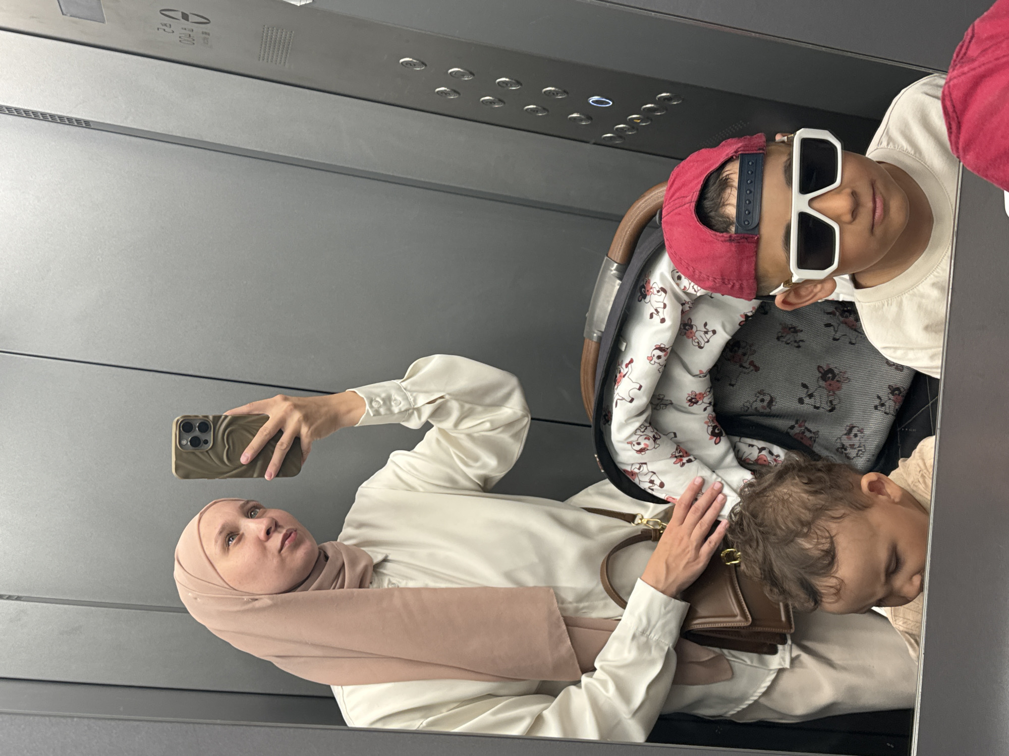 A reflection shows a woman and two children in what appears to be an elevator. The woman wears a hijab and white blouse, and is holding a smartphone. One child wears sunglasses and a baseball cap, sitting in a stroller. The other child is asleep or resting, with their head reclined. The interior of the elevator appears to be modern with grey walls and a control panel visible at the top. Without additional information or access to specific metadata, the precise location within Chaikovsky, Russia cannot be confirmed solely from this image.