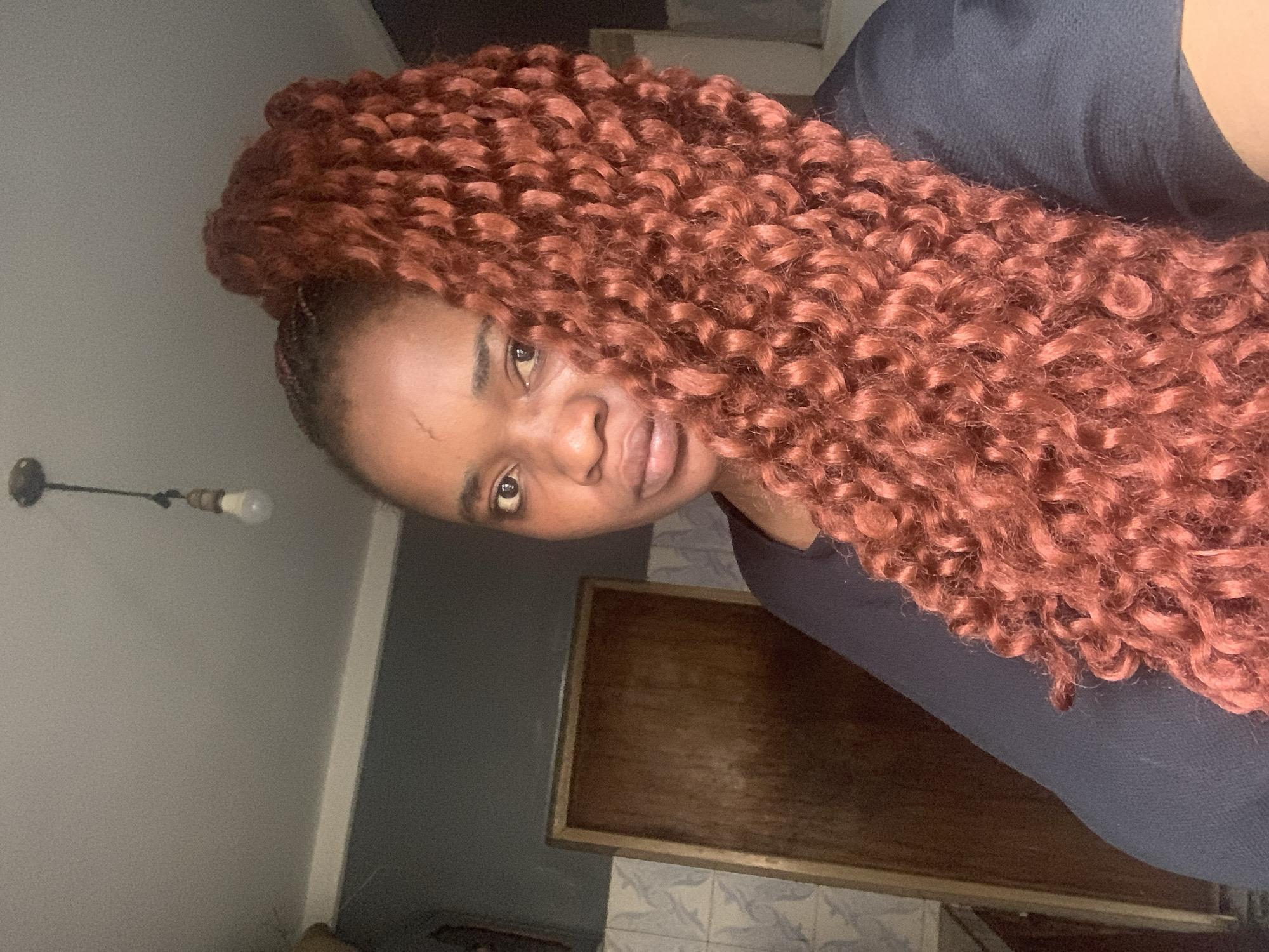 A portrait-style image captures a young Black woman in an indoor setting, consistent with residential interiors in Jos, Nigeria. She is positioned slightly off-center, with her gaze directed towards the camera, exhibiting a calm and neutral expression.
She has long, voluminous, reddish-brown or auburn hair styled in tight, bouncy curls, likely a protective style such as crochet braids. The hair cascades over her right shoulder and fills a significant portion of the right side of the frame. She is wearing a dark, simple top, possibly black or navy.
The background consists of plain light-grey walls. To her upper left, a simple light fixture with a bare, frosted incandescent bulb is mounted on the wall. To her right and lower right, a dark brown wooden frame, possibly for a mirror or artwork, is visible against a wall section covered with light-colored tiles featuring a subtle blueish-grey pattern. The overall lighting is soft and appears to be artificial, illuminating her face and hair. No text is visible in the image. The woman is posing for a casual photograph or selfie.