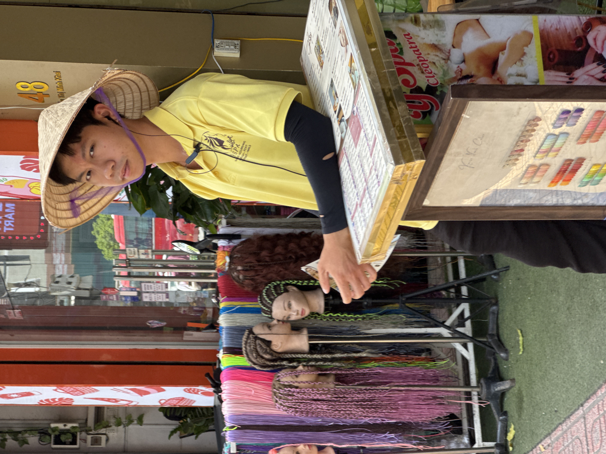 A young man wearing a conical hat and a yellow polo shirt is standing behind a display of colorful wigs and artificial hair. He appears to be a vendor. The display includes mannequins with braided hairstyles in various colors, such as purple, gray, and neon green. Behind the wigs, a rack of colorful hair strands is visible. The man is holding a small menu or display board. In the background, there is a building with signage, including the number "48" and text in Vietnamese. Part of a red bus is visible through a glass door. The scene appears to be on a city street, possibly in a market or commercial area. The ground is covered with artificial turf. The overall impression is a street vendor operation, likely selling hair accessories or beauty services in Nha Trang, Vietnam.