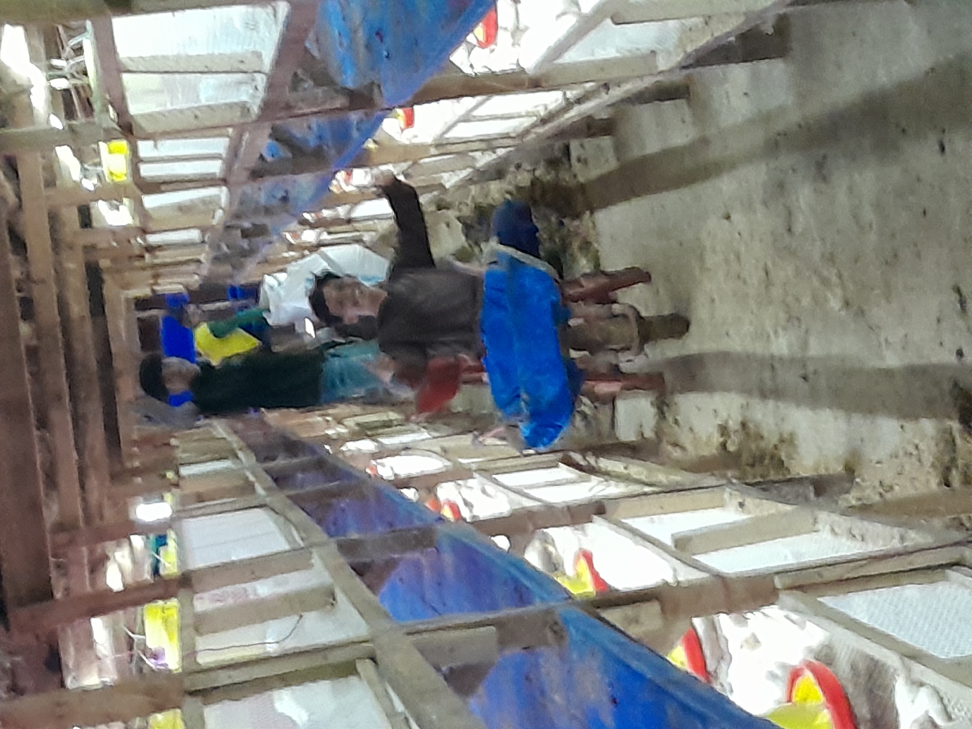 In Labuhan Ratu Lima, Indonesia, the image depicts a dimly lit, long poultry farm structure. The scene is dominated by wooden beams and shelves that form rows of cages or enclosures. Several individuals are visible within this environment. In the foreground, a person wearing a dark shirt and blue pants is seated on a structure that appears to be a stool or a repurposed piece of equipment, covered with a blue tarp. This individual is smiling and gesturing with their hands. Further down the structure, another person in a dark shirt and blue jeans stands, holding a white bag. The floor of the farm is dusty and appears to have accumulated debris. Red and yellow feeders are visible within the enclosures. The overall impression is one of a working poultry farm, possibly undergoing maintenance or cleaning, with people engaged in activities within.