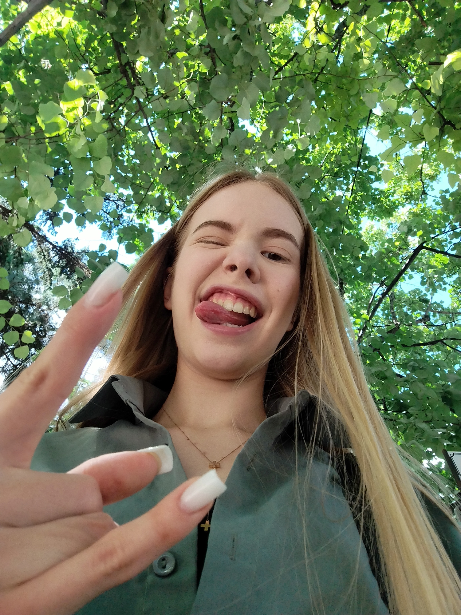 A young woman with long blonde hair is taking a selfie outdoors. She is wearing a green shirt and a necklace with a cross pendant. She is winking and sticking her tongue out, with her right hand raised in a peace sign. The background is filled with green leaves and blue sky, suggesting the photo was taken under a tree on a sunny day. The location is unknown.