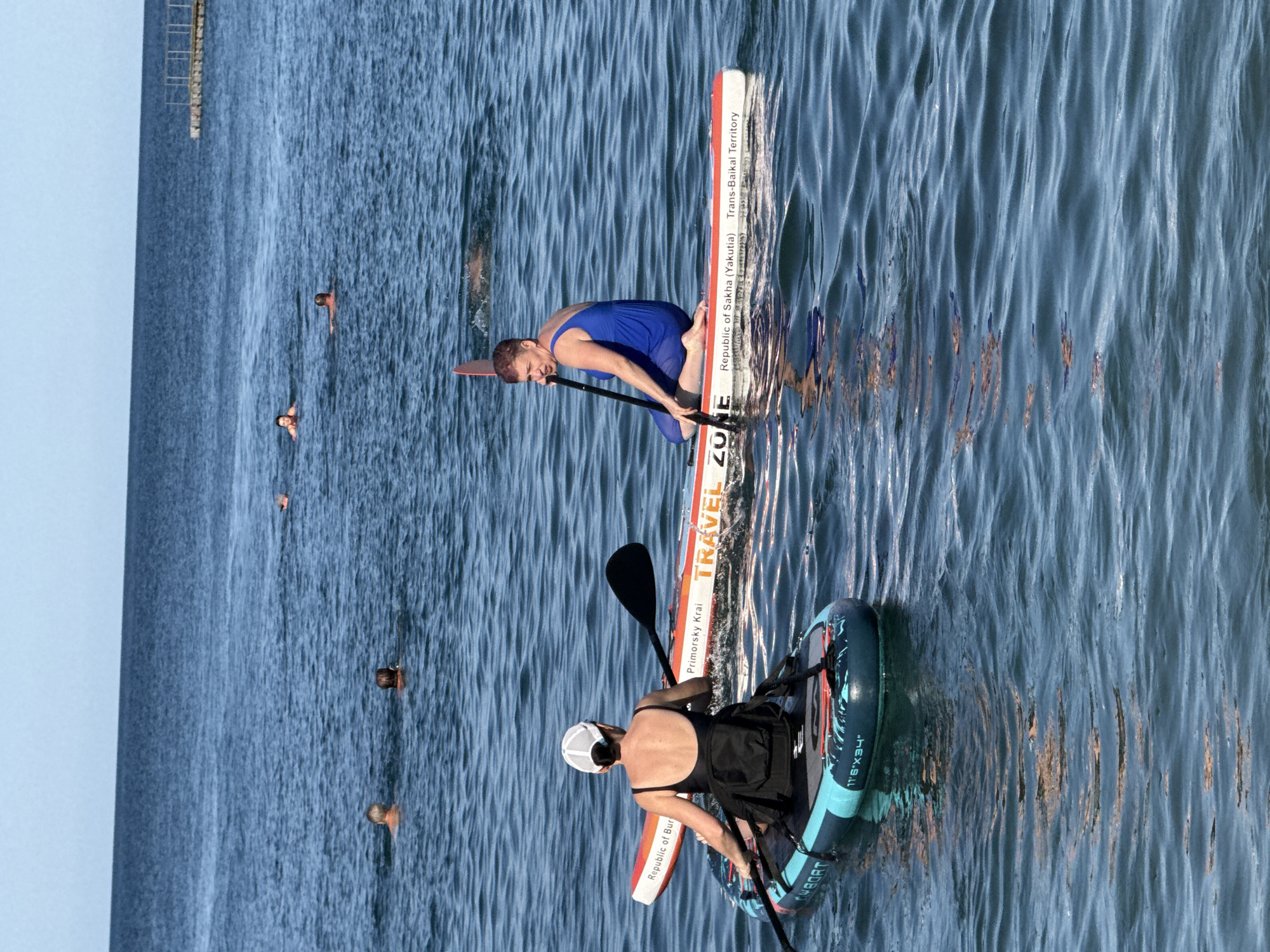 In the coastal waters of Partenit, Ukraine, on a clear, sunny day, two individuals are engaged in paddleboarding. One person, seen from behind, is seated on a paddleboard, wearing a black swimsuit and a white cap. They are holding a paddle, poised to move through the water. Another person, facing left and wearing a blue one-piece swimsuit, is kneeling on a paddleboard, also holding a paddle. This paddleboard has text visible on its side, including "TRAVEL ZONE," "Primorsky Krai," "Republic of Sakha (Yakutia)," and "Trans-Baikal Territory." In the background, several other people are swimming and paddling in the calm, blue sea, with a distant pier structure visible on the horizon. The water is gently rippled, reflecting the clear sky.