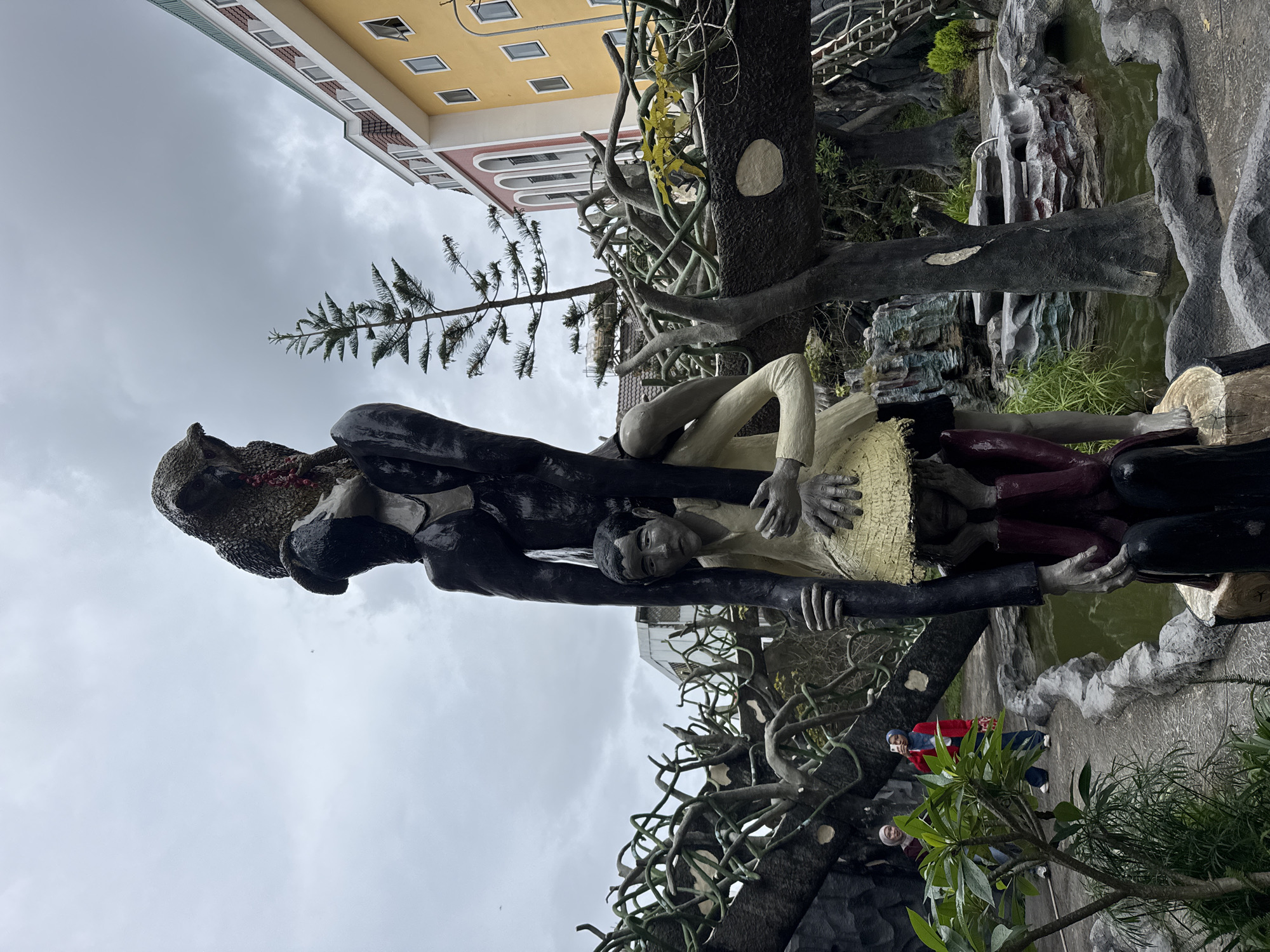 The image displays a complex, multi-figure sculpture set within a fantastical, artificial landscape, under an overcast sky. The primary subject is a large, dark, possibly bear-like or monster-like figure with reddish details on its face, acting as a central anchor. Clinging to this dark figure are two human-like figures. One, wearing a light-colored (cream or yellow) top and dark trousers, looks towards the viewer with an open expression while nestled within the larger figure's embrace. Another dark, slender figure also appears to be part of this intertwined group, possibly supporting or holding the lighter-clad figure. The sculpture itself integrates with gnarled, tree-like structures and branches that form part of the immediate environment.
The scene is an outdoor art installation, likely part of a themed garden or attraction. To the right of the sculpture, a body of water, possibly a pond or stream, is visible with artificial rock formations along its banks. In the background, a multi-story building with a distinctive yellow facade, white trim, and numerous rectangular windows stands out against the cloudy sky. A coniferous tree branch extends into the upper left of the frame. Several other visitors are visible in the background, near the water, observing the surroundings; one person wears a red jacket, and another has a light-colored head covering.
The weather is cloudy, with a uniform grey sky suggesting an overcast day. The dramatic and somewhat eerie nature of the sculpture, combined with the unique architecture in the background, points to a creative and artistic environment, consistent with tourist attractions found in Da Lat, Vietnam. No discernible text is visible in the image.
