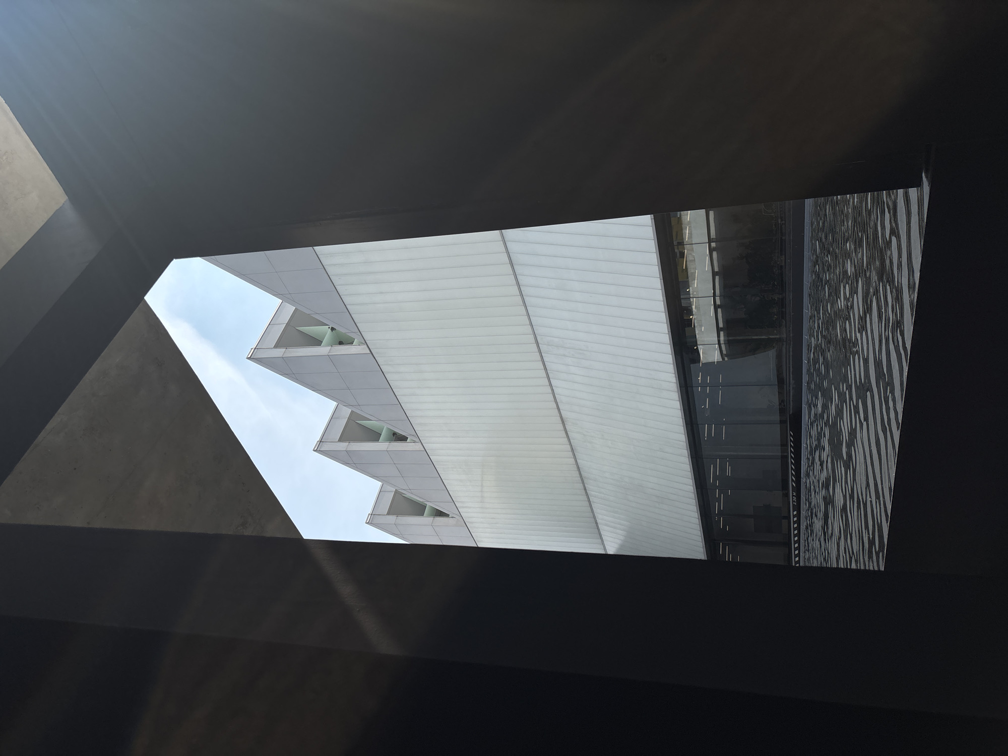 A low-angle shot taken from a confined space captures a modern building with a distinctive sawtooth roofline. The building has a white facade, and each peak of the sawtooth roof features a small triangular window. The view of the building is framed by dark, angular concrete structures, suggesting a close-up perspective from within a building or under an overpass. To the right of the white facade, there's a glimpse of a more conventional building with glass windows and a decorative panel, which is adorned with an abstract design. The sky, visible only in a small section above the sawtooth roof, is pale blue. Based on the architecture and design elements, this image is likely taken in Bangkok, Thailand, which is known for its contemporary buildings.