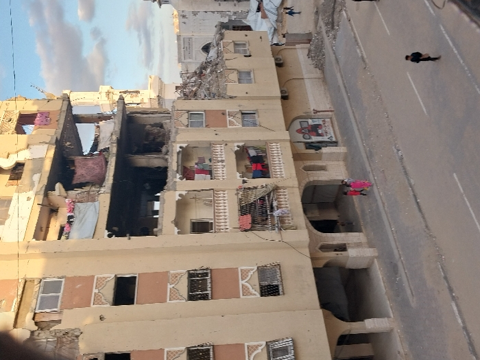 This image depicts a damaged multi-story building in Khan Yunis, Palestinian Territory, under daylight conditions. The beige-colored structure, featuring decorative window and balcony elements, exhibits significant structural damage across its upper levels, particularly from the third to the sixth visible floors. Sections of exterior walls are missing, revealing exposed interiors, rebar, and debris within the collapsed portions. Shattered or absent windows are visible on multiple floors. Laundry is observed hanging on balconies of the second, third, and fourth visible floors. On the third floor balcony, one individual in a light blue shirt is present. The ground floor features arched entryways leading to a recessed area, and a red and black graphic sign is affixed to the wall to the right of these arches.
In the foreground, a paved street runs parallel to the building. It features white solid and dashed lane markings and is dusted with debris. Three children are walking on the street near the building's ground-floor arches; two are wearing bright pink/red clothing, and one is in darker attire. Further down the street to the right, a solitary figure in dark clothing walks away from the viewer. Along the sidewalk and at the base of the damaged building, piles of rubble and debris are scattered.
In the background, to the right of the main building, a large, light-colored structure resembling a mosque with a prominent minaret is visible, also showing signs of damage or wear. Adjacent to this background building, an accumulation of rubble and what appear to be temporary shelters or tents are present, with several small figures observable in that distant area. The sky is partly cloudy with patches of blue.