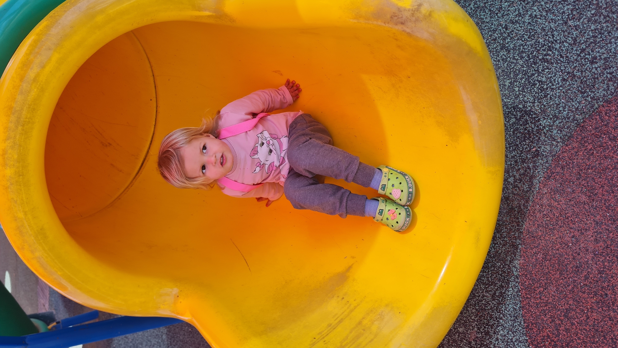 A young child is inside a yellow tunnel slide. The child, with blonde hair, is wearing a pink shirt with a cat design, gray pants, and green shoes with a duck pattern. The child is lying on their back and looking up at the camera. The slide appears to be part of a playground with colorful surfacing next to the slide. The location is Lachish Regional Council, Israel.