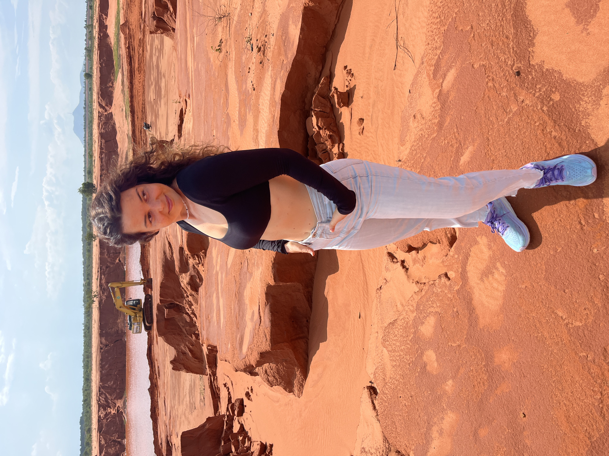 A young woman with curly brown hair stands in a red sand dune landscape in Mui Ne, Vietnam. She is wearing a black long-sleeved crop top and light-wash jeans, with her left hand in her pocket. Her right hand is by her side. She is wearing blue athletic shoes with purple laces. In the background, a yellow excavator is visible near a body of pink water. The terrain consists of reddish-orange sand with eroded formations. The sky is blue with scattered clouds.