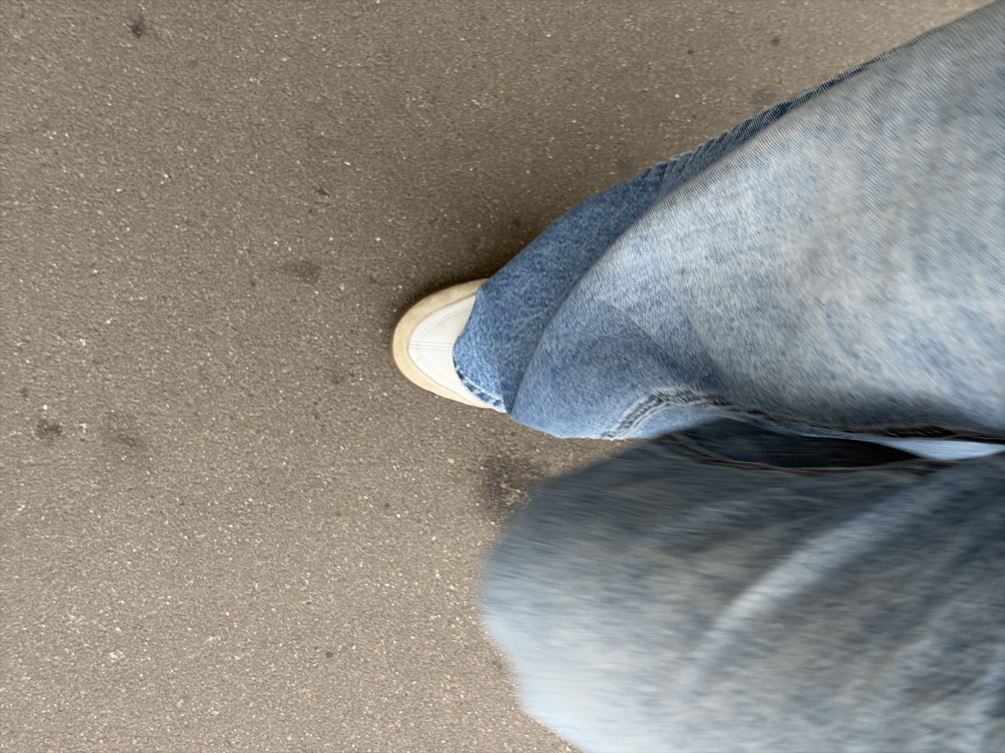 The image shows a first-person perspective of someone walking on asphalt. The person is wearing light-wash denim jeans and white sneakers. The left leg and foot are blurred due to motion, while the right leg and foot are in focus, showing the details of the denim and the white toe of the shoe. The ground is a textured asphalt surface with small, light-colored pebbles. No other individuals or structures are visible. The scene suggests an outdoor, urban environment.